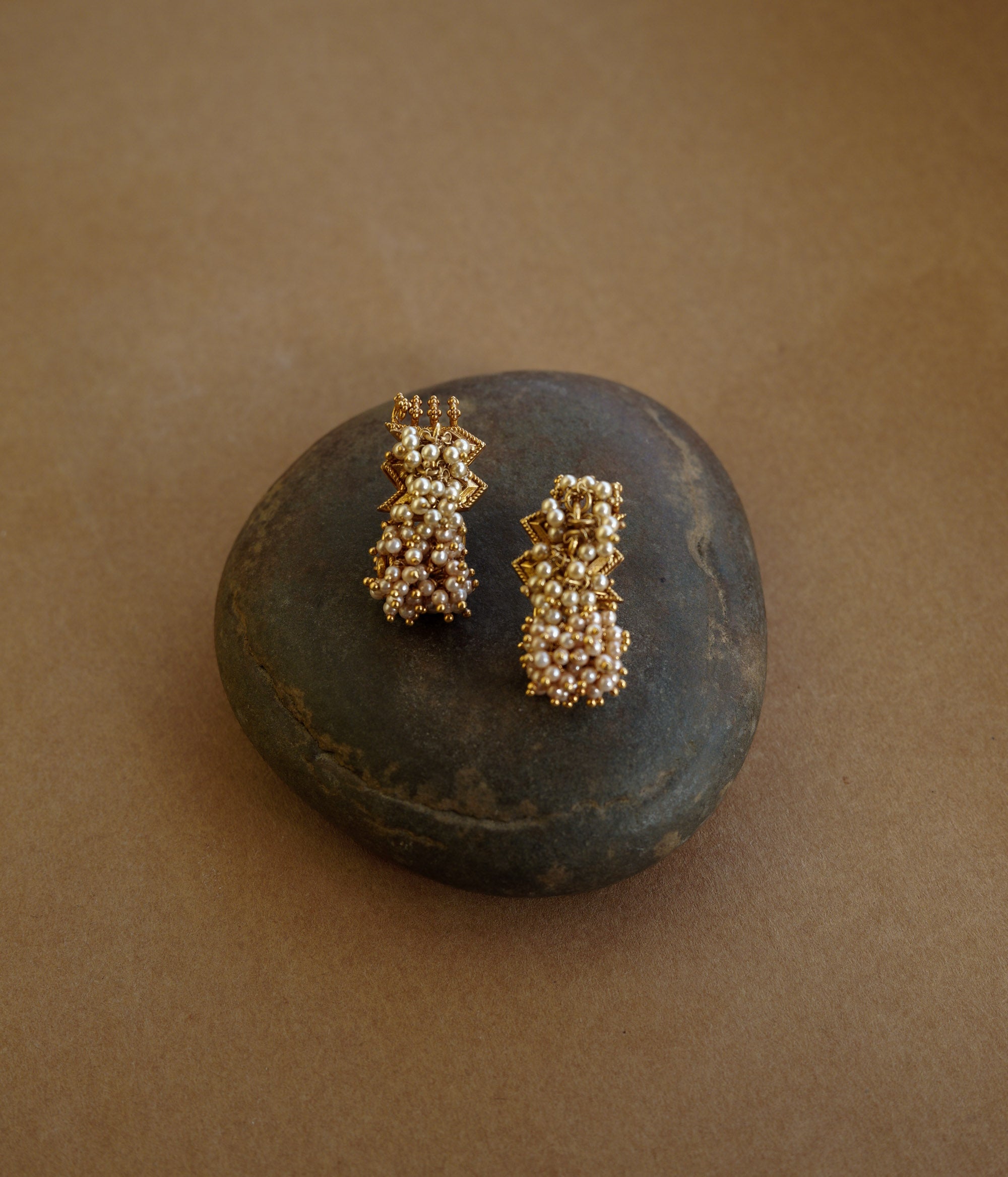 Gold earrings with a jagged, star-like motif at the top and a cluster of golden-hued beads/pearls dangling below. The earrings are on a textured brown surface.