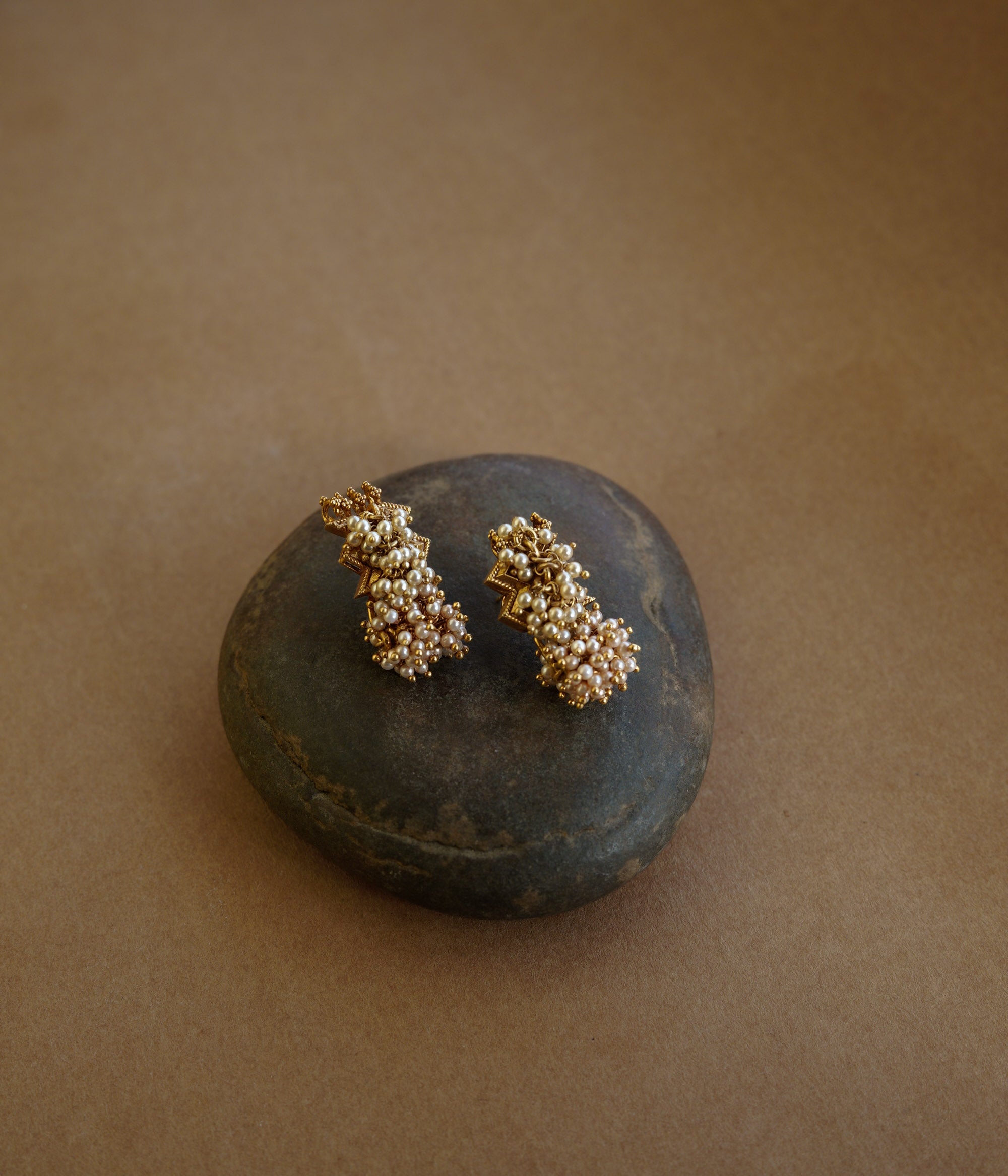 Gold earrings with a jagged, star-like motif at the top and a cluster of golden-hued beads/pearls dangling below. The earrings are on a textured brown surface.