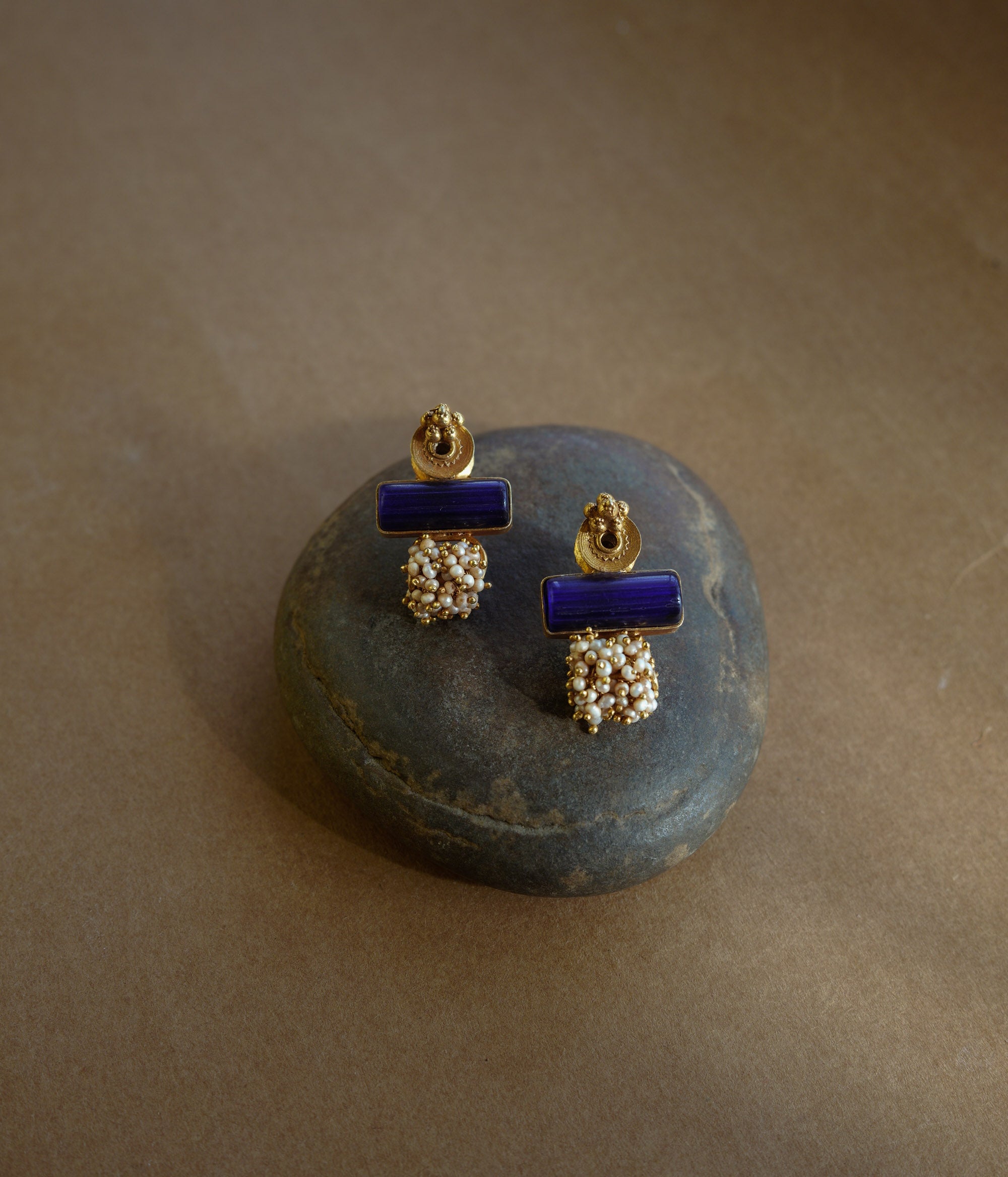 Gold earrings with a rectangular blue stone at the top and a cluster of small white pearls dangling below, resting on a textured brown surface.