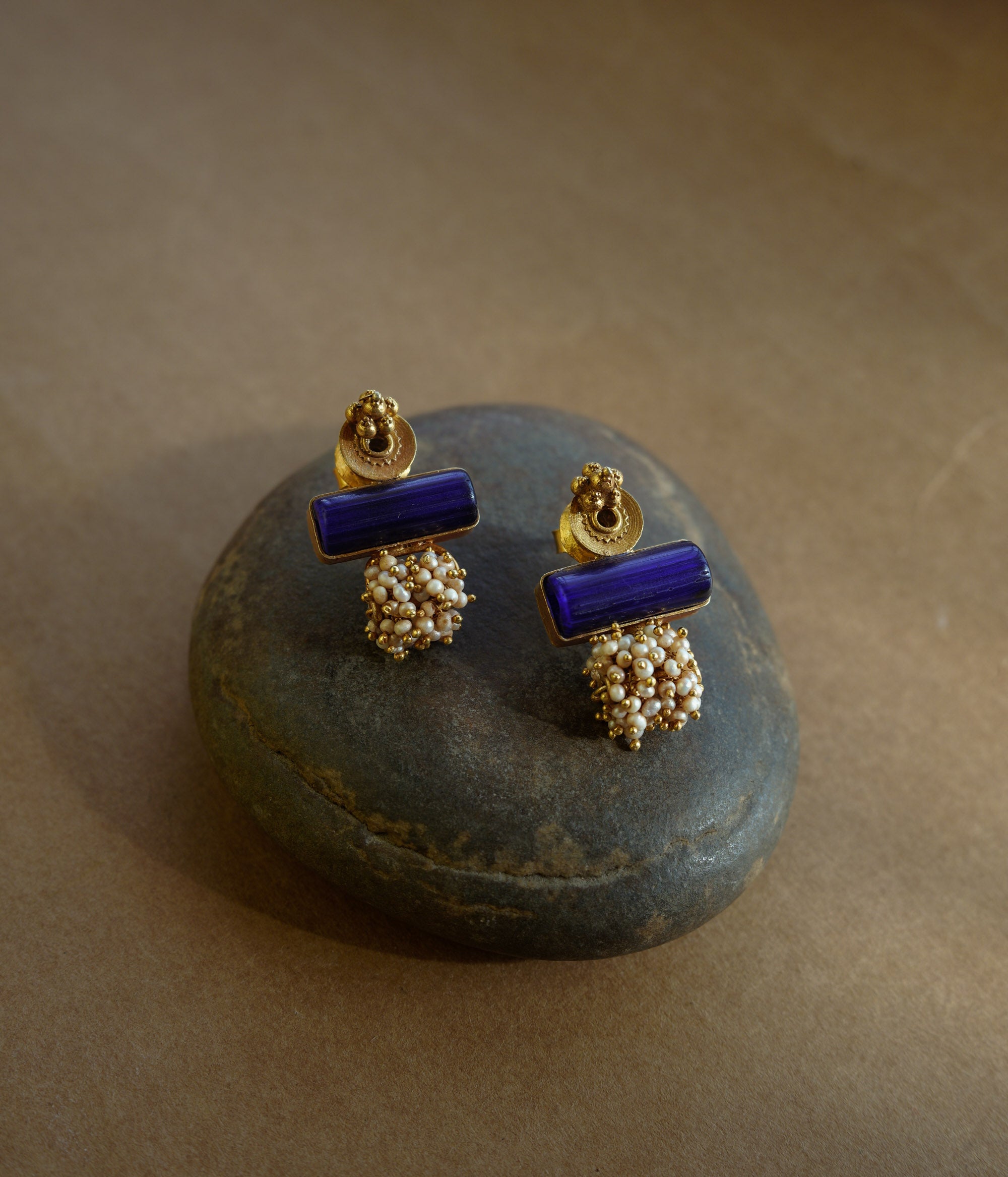 Gold earrings with a rectangular blue stone at the top and a cluster of small white pearls dangling below, resting on a textured brown surface.