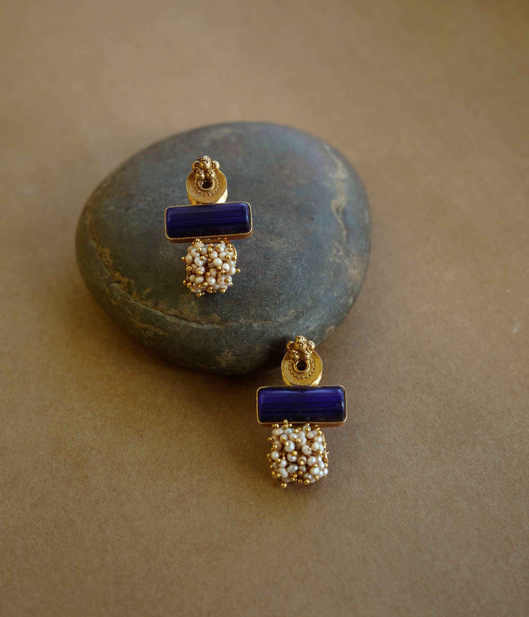 Gold earrings with a rectangular blue stone at the top and a cluster of small white pearls dangling below, resting on a textured brown surface.