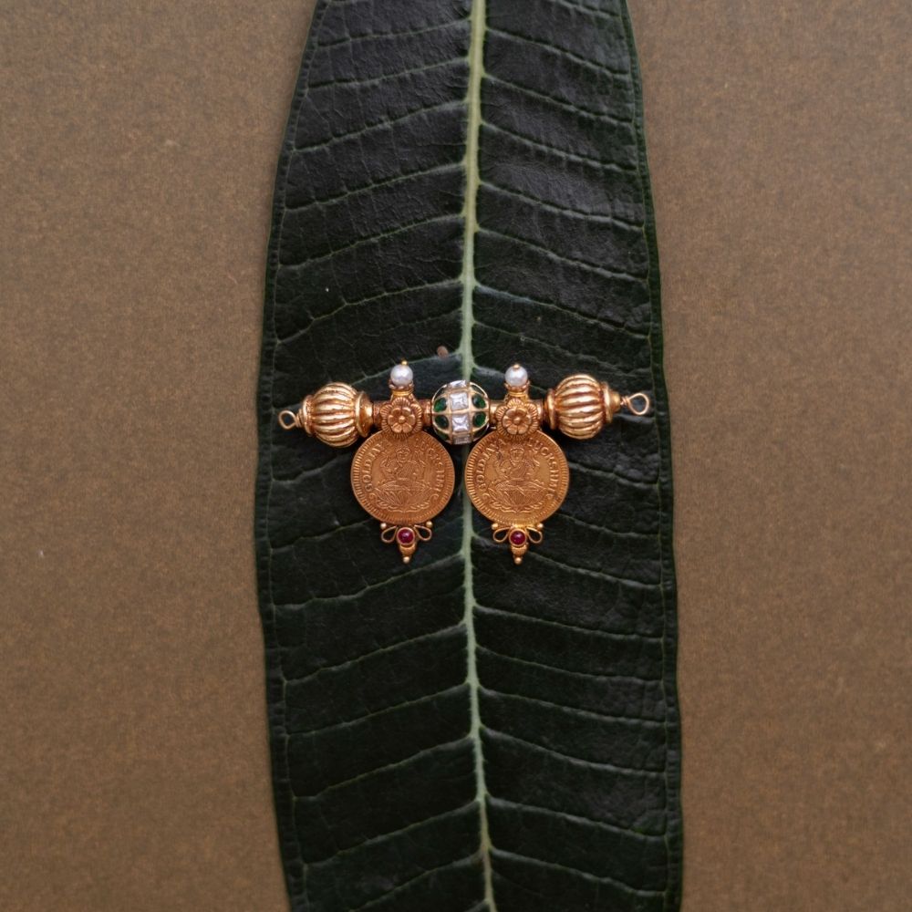Antique gold coin pendant with red spinel, red and green kundan, and pearls, displayed on a long green leaf.
