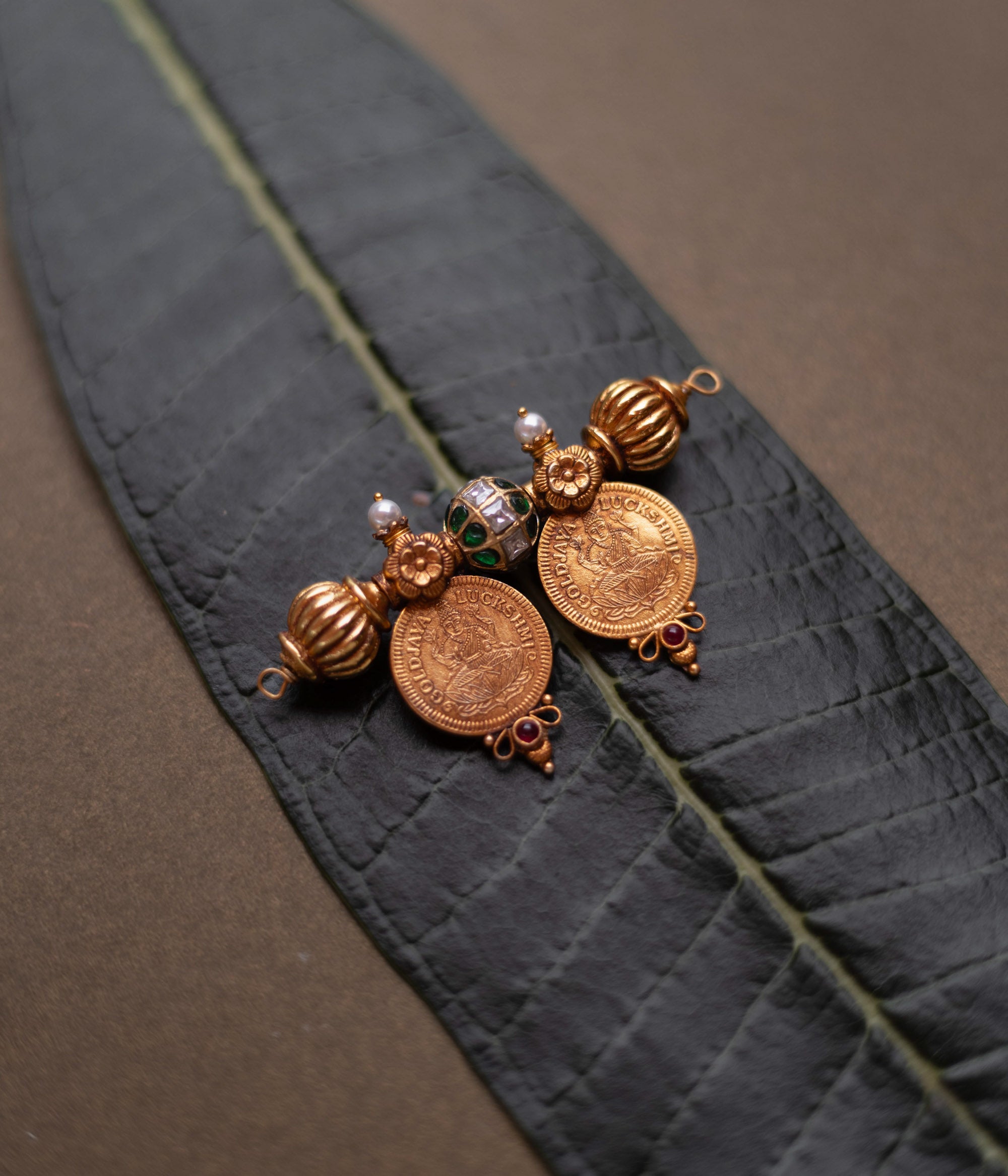Antique gold coin pendant with red spinel, red and green kundan, and pearls, displayed on a long green leaf.