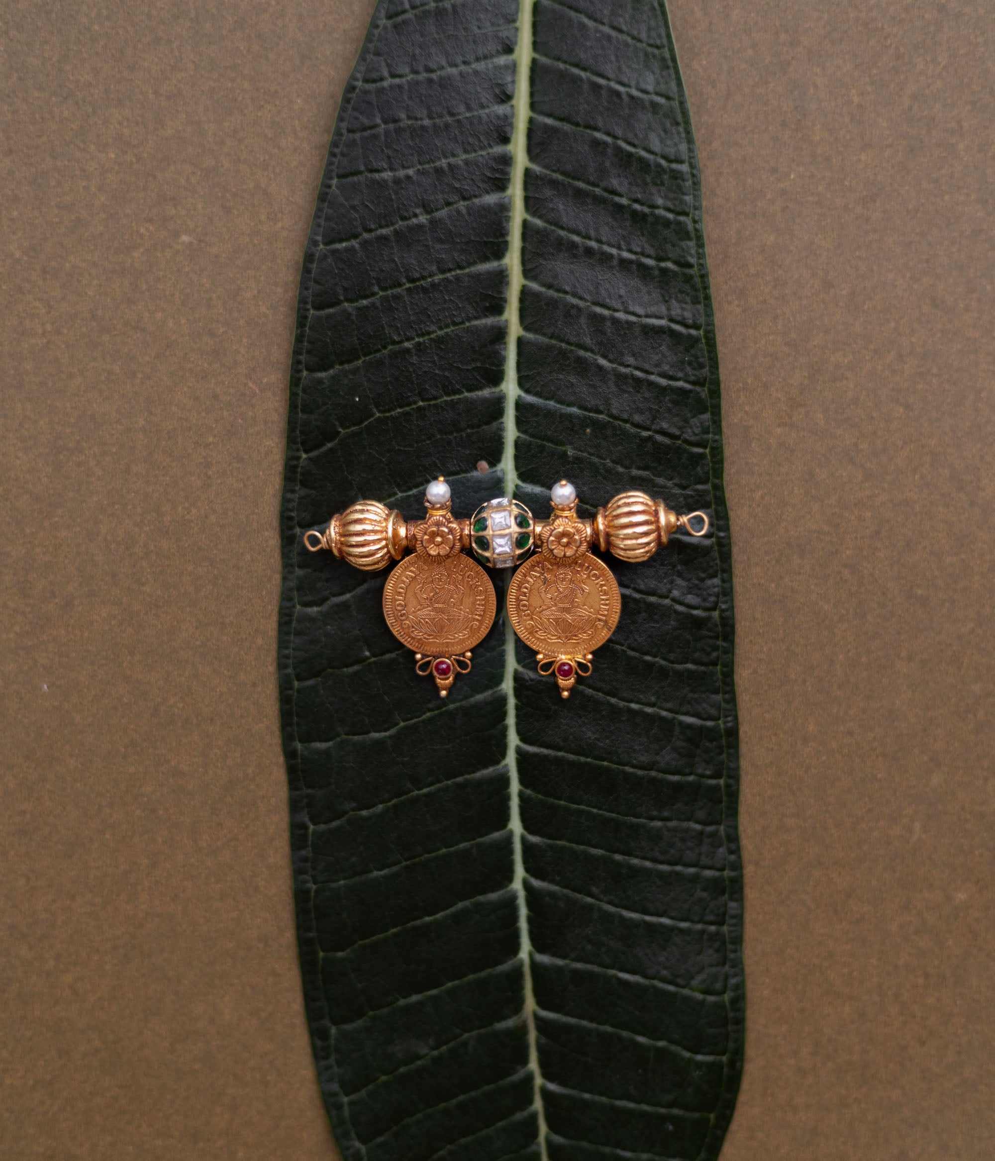 Antique gold coin pendant with red spinel, red and green kundan, and pearls, displayed on a long green leaf.