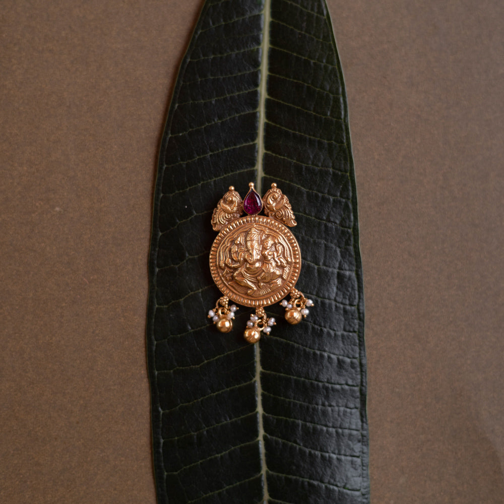 Antique gold Ganesha pendant with red spinel stone and pearl drops, on a dark green leaf.