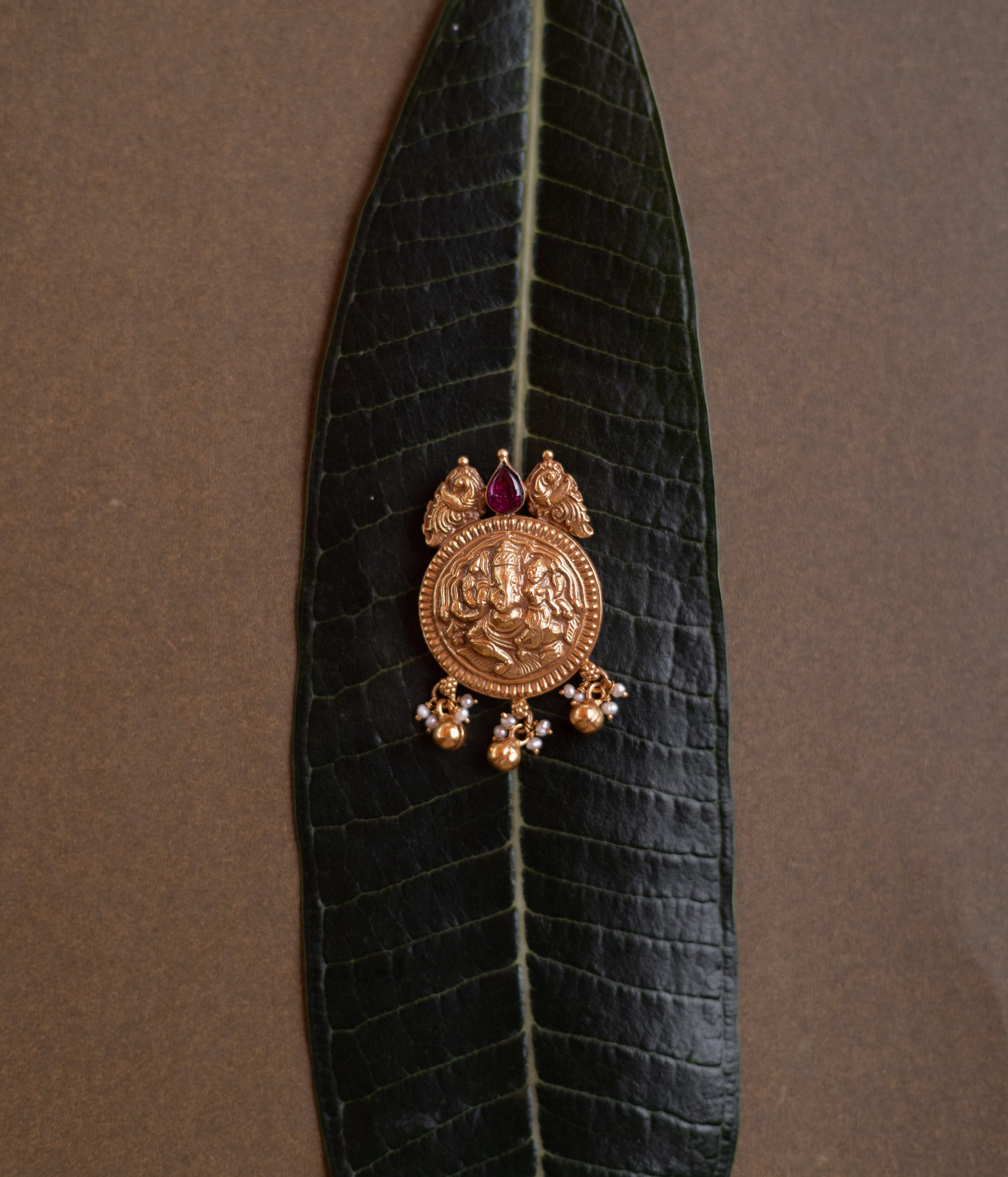 Antique gold Ganesha pendant with red spinel stone and pearl drops, on a dark green leaf.
