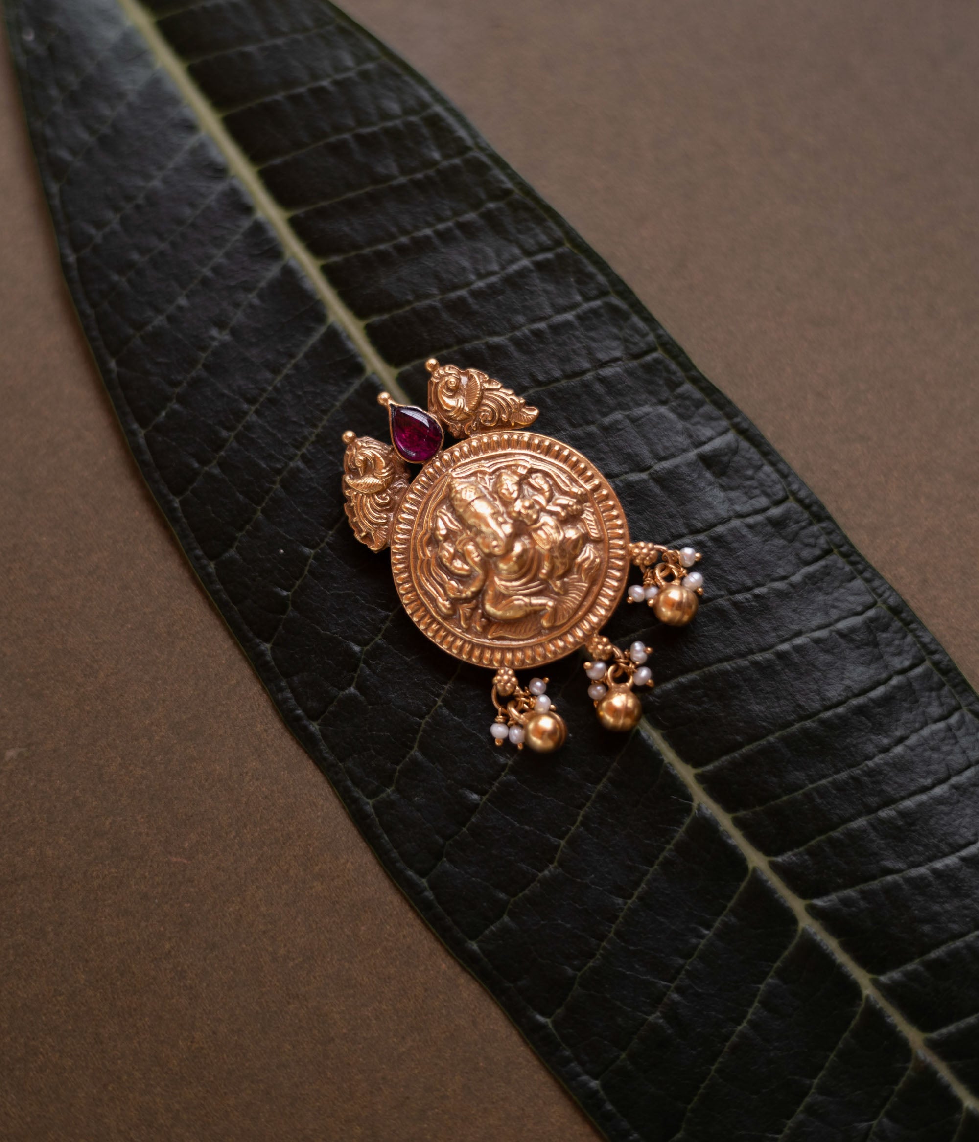 Antique gold Ganesha pendant with red spinel stone and pearl drops, on a dark green leaf.