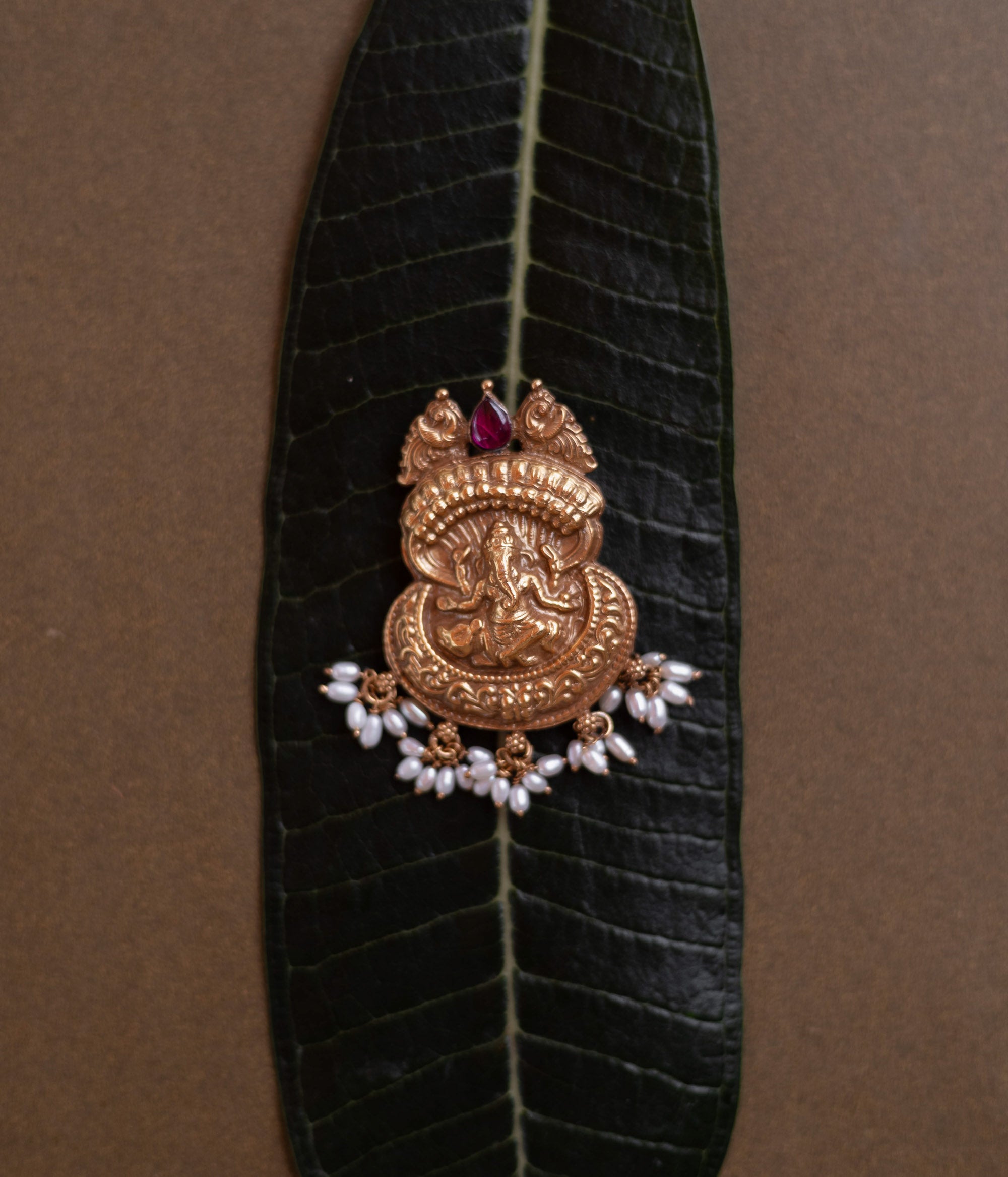 Antique gold Ganesha pendant with a conch-like top, red spinel, and pearl drops, on a dark green leaf.