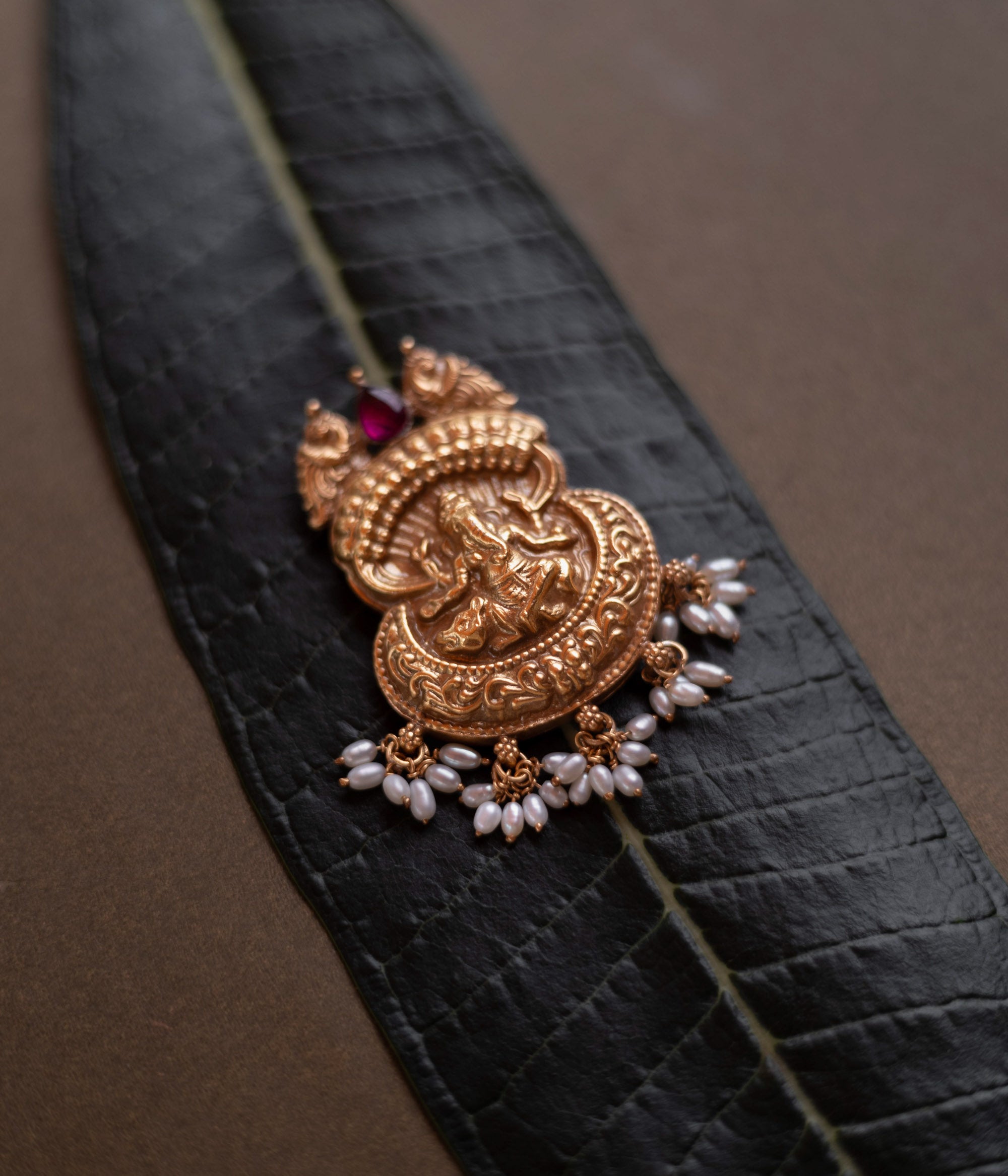 Antique gold Ganesha pendant with a conch-like top, red spinel, and pearl drops, on a dark green leaf.