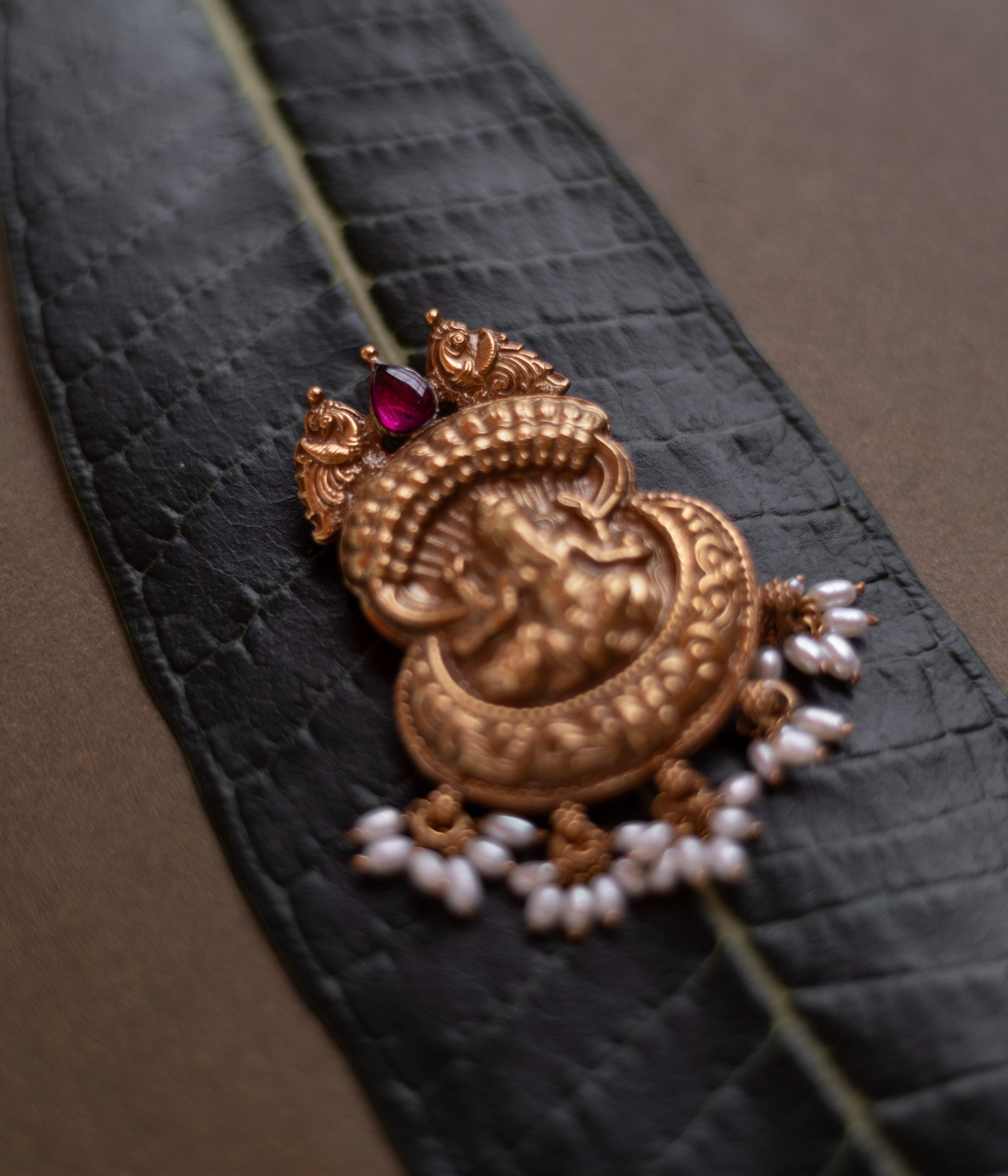 Antique gold Ganesha pendant with a conch-like top, red spinel, and pearl drops, on a dark green leaf.