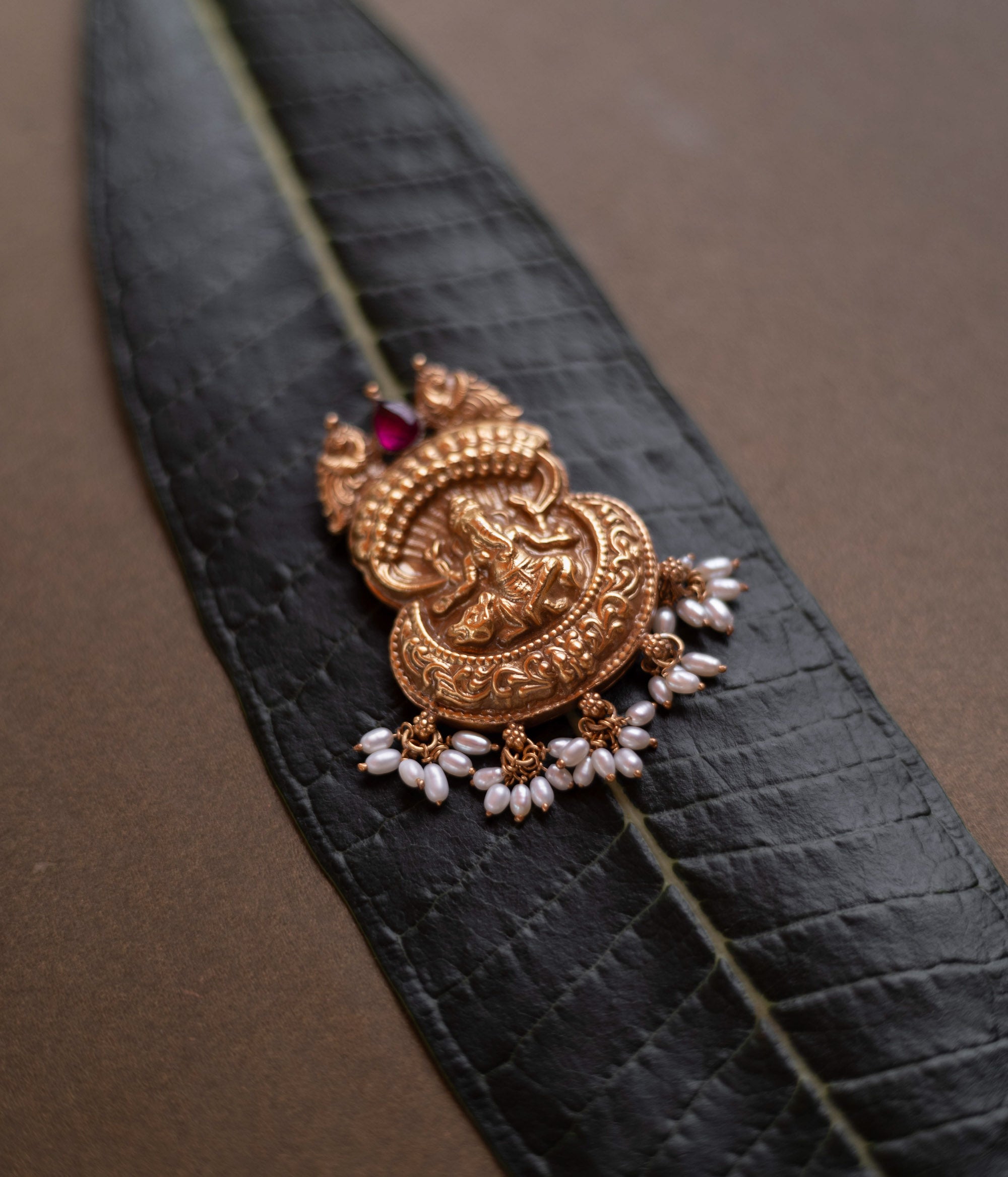 Antique gold Ganesha pendant with a conch-like top, red spinel, and pearl drops, on a dark green leaf.