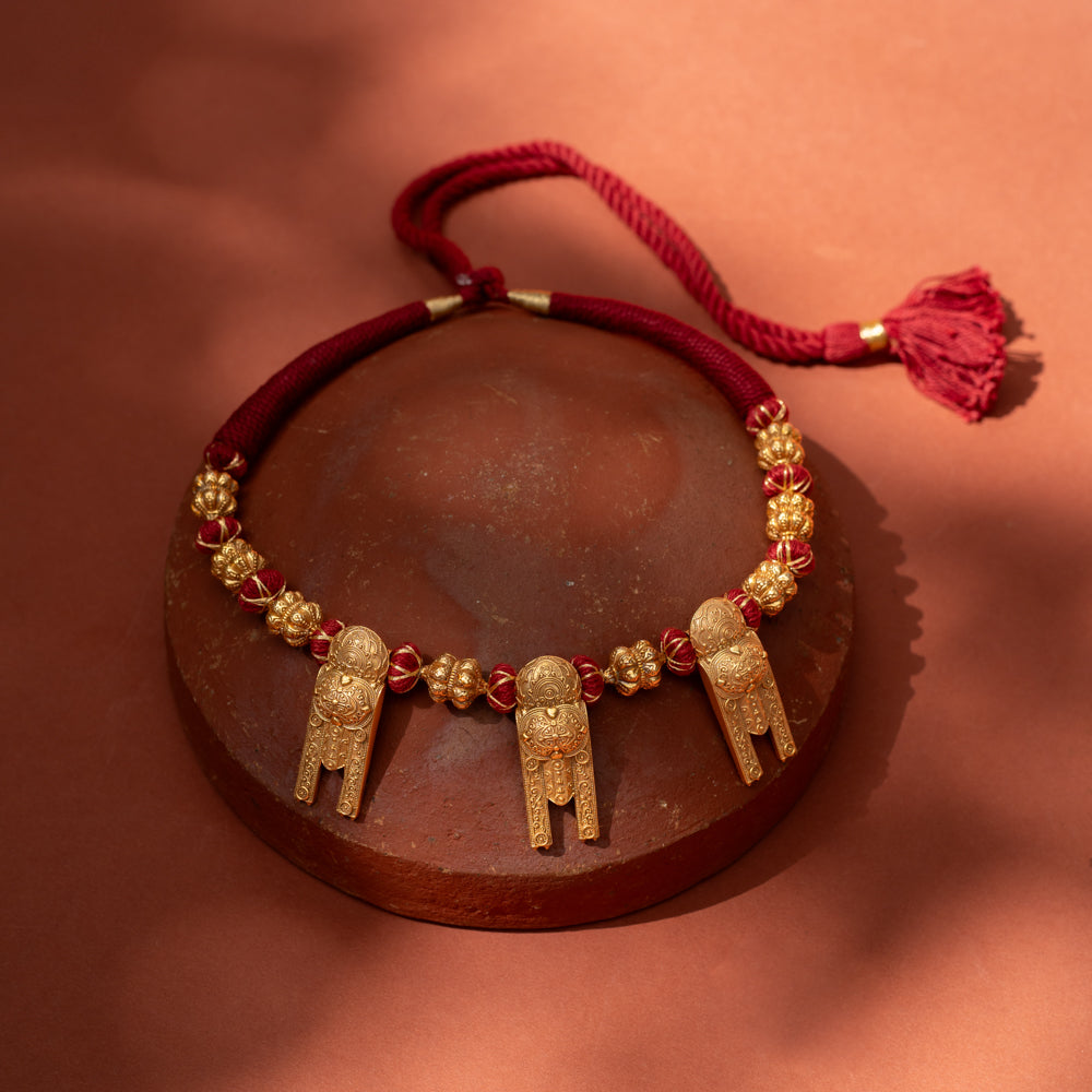 A short, bold necklace with a red braided cord. Three ornate, rectangular gold tribal pendants hang from it, interspersed with golden textured beads, resting on a terracotta surface.