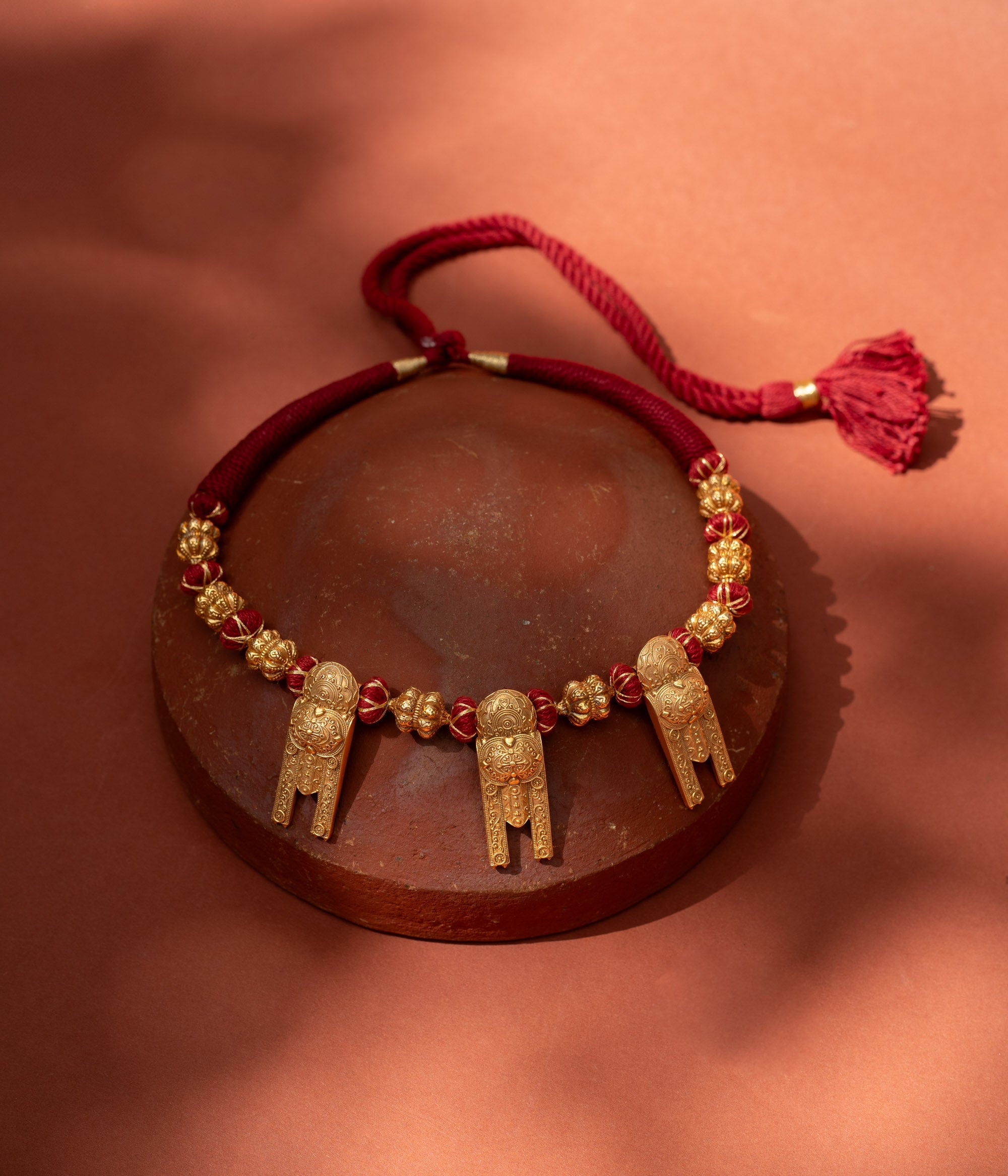 A short, bold necklace with a red braided cord. Three ornate, rectangular gold tribal pendants hang from it, interspersed with golden textured beads, resting on a terracotta surface.