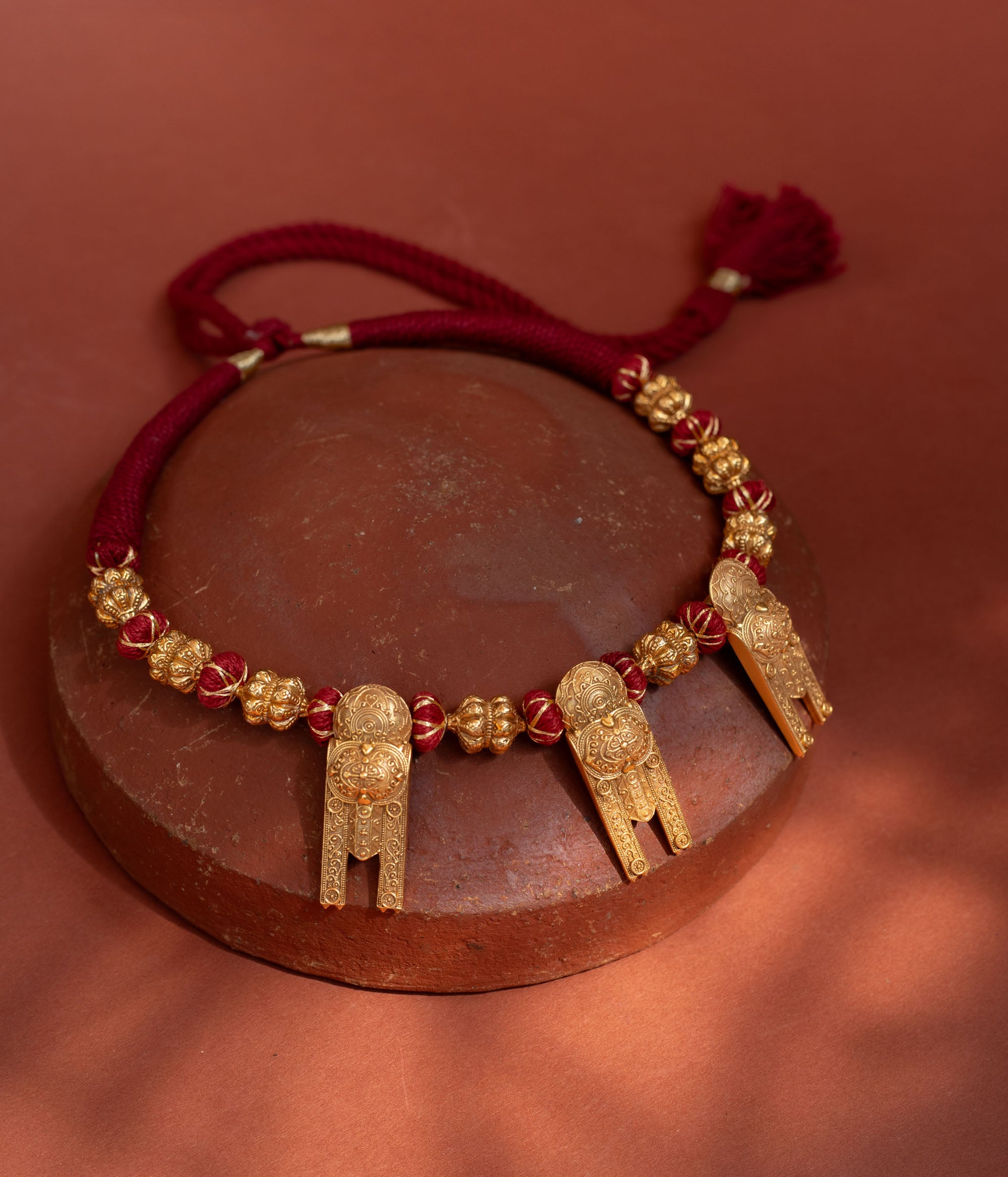 A short, bold necklace with a red braided cord. Three ornate, rectangular gold tribal pendants hang from it, interspersed with golden textured beads, resting on a terracotta surface.