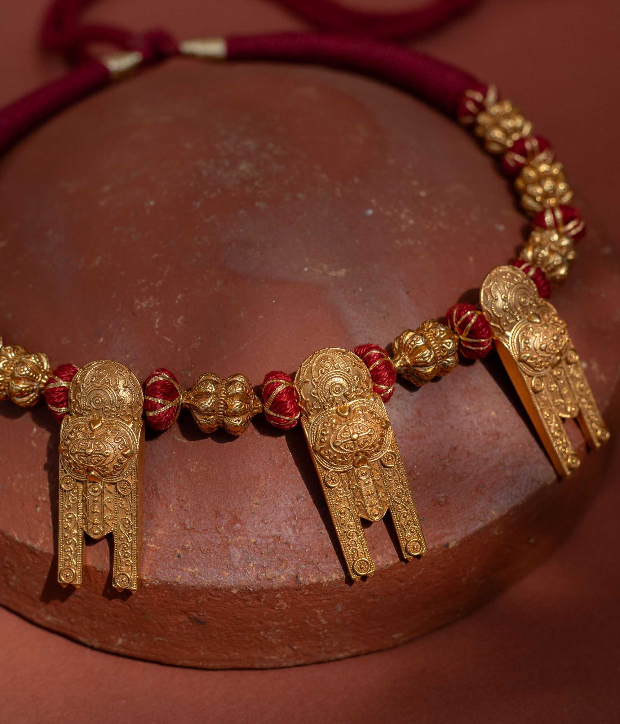 A short, bold necklace with a red braided cord. Three ornate, rectangular gold tribal pendants hang from it, interspersed with golden textured beads, resting on a terracotta surface.