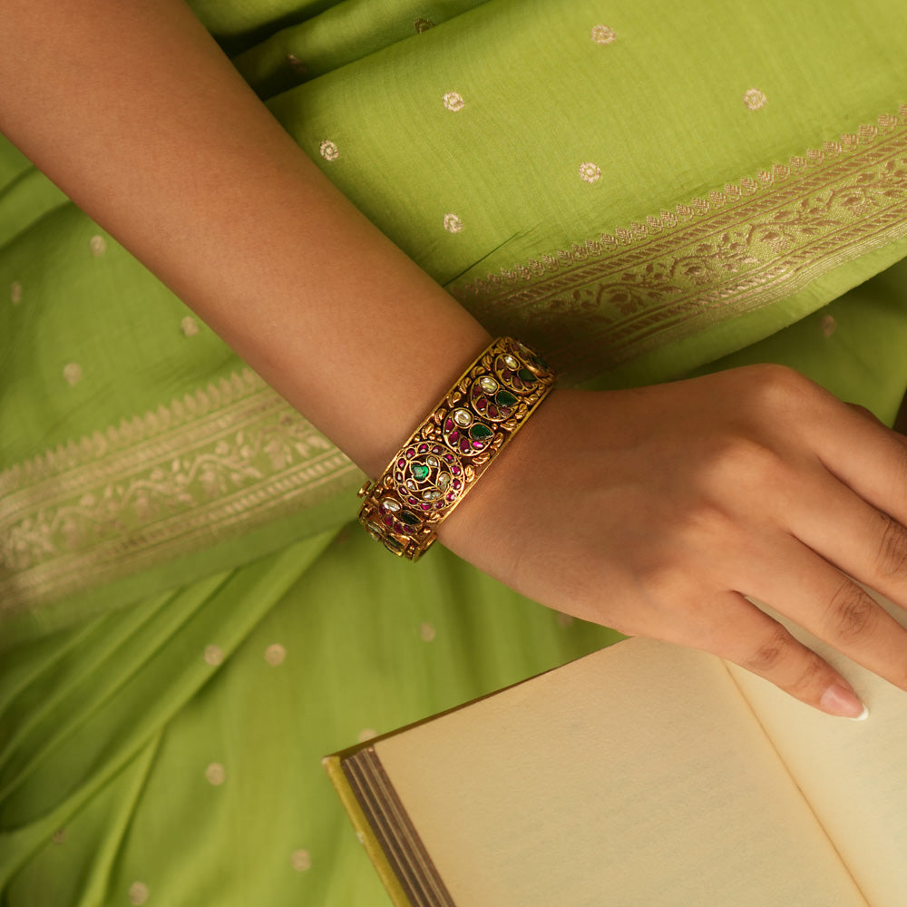 A stunning bangle featuring a peacock motif, adorned with red, white, and green Kundan stones. The vibrant hues of the Kundan stones gleam, embodying royal opulence. A masterpiece of intricate craftsmanship, this bangle exudes timeless allure.