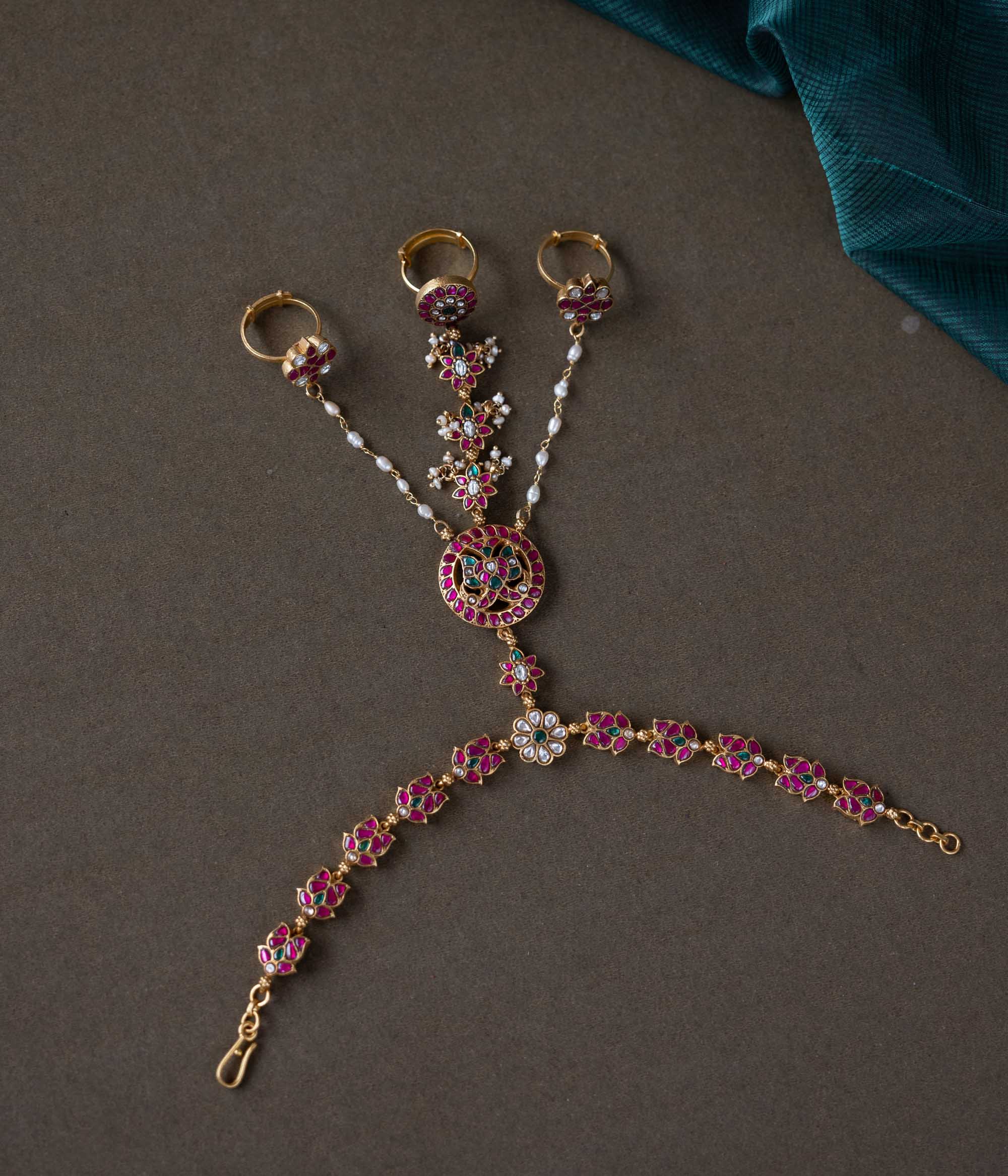 Handmade 92.5 sterling silver and 22ct gold-plated Trishika Haathphool, adorned with white, red, and green Kundan stones and pearls. An intricate masterpiece of elegance and tradition, weaving royal charm with delicate floral accents.
