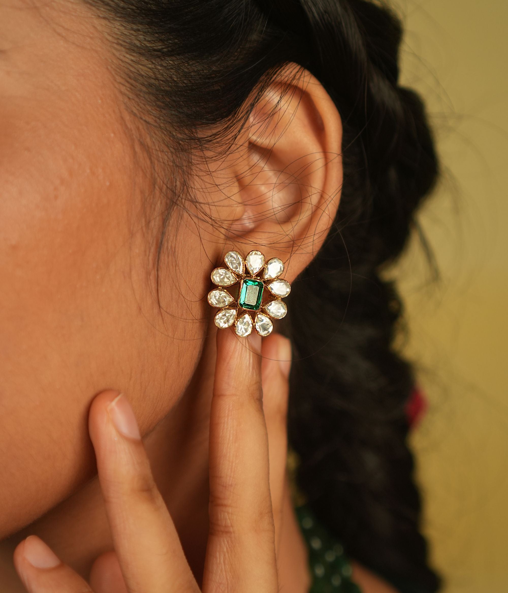 Amidst the vibrant charm of a bustling bazaar, this minimal floral stud with a radiant green cut stone at its heart, encircled by shimmering polki stones, whispers timeless elegance.