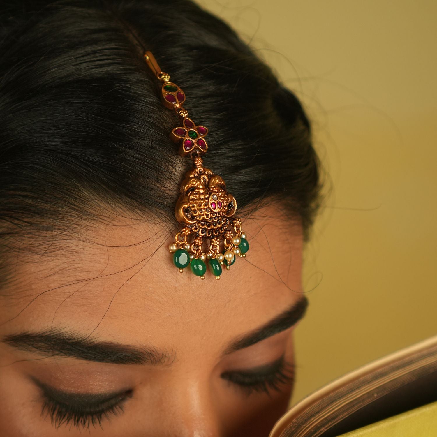 In the serene elegance of a heritage palace, this maangtika gleams with red, green, and white Kundan stones, complemented by lustrous pearls. Its temple-inspired design pays a timeless tribute to regal splendor and tradition.