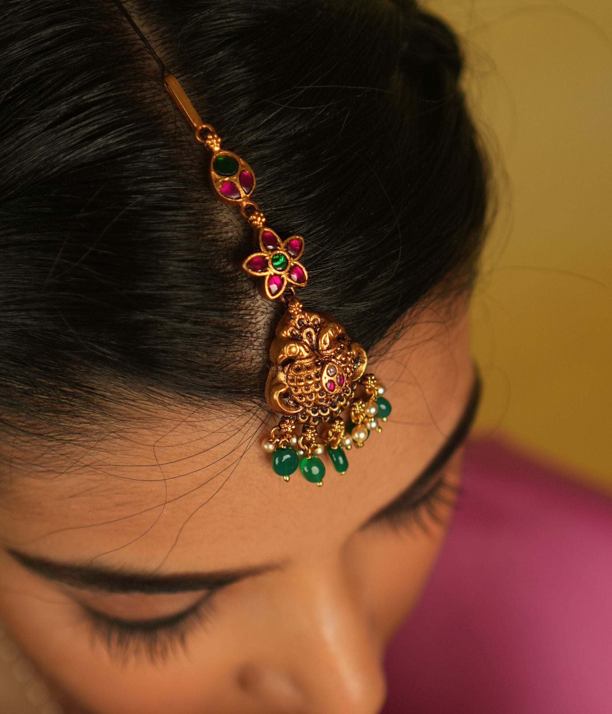 In the serene elegance of a heritage palace, this maangtika gleams with red, green, and white Kundan stones, complemented by lustrous pearls. Its temple-inspired design pays a timeless tribute to regal splendor and tradition.