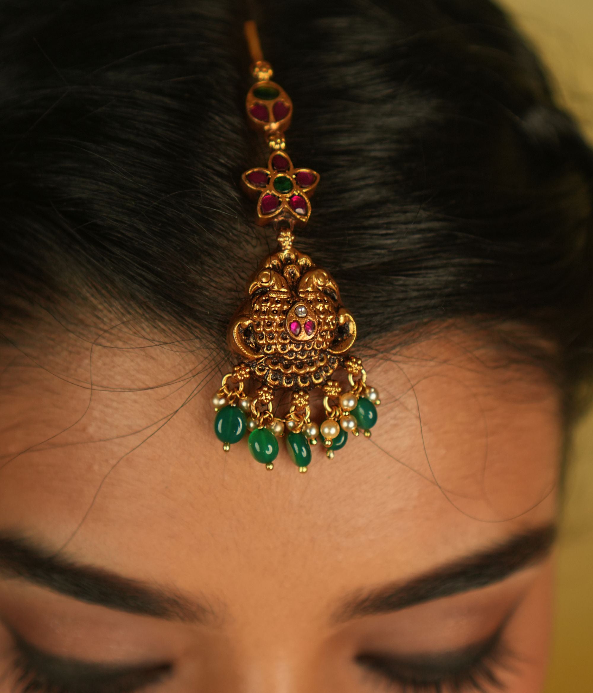 In the serene elegance of a heritage palace, this maangtika gleams with red, green, and white Kundan stones, complemented by lustrous pearls. Its temple-inspired design pays a timeless tribute to regal splendor and tradition.