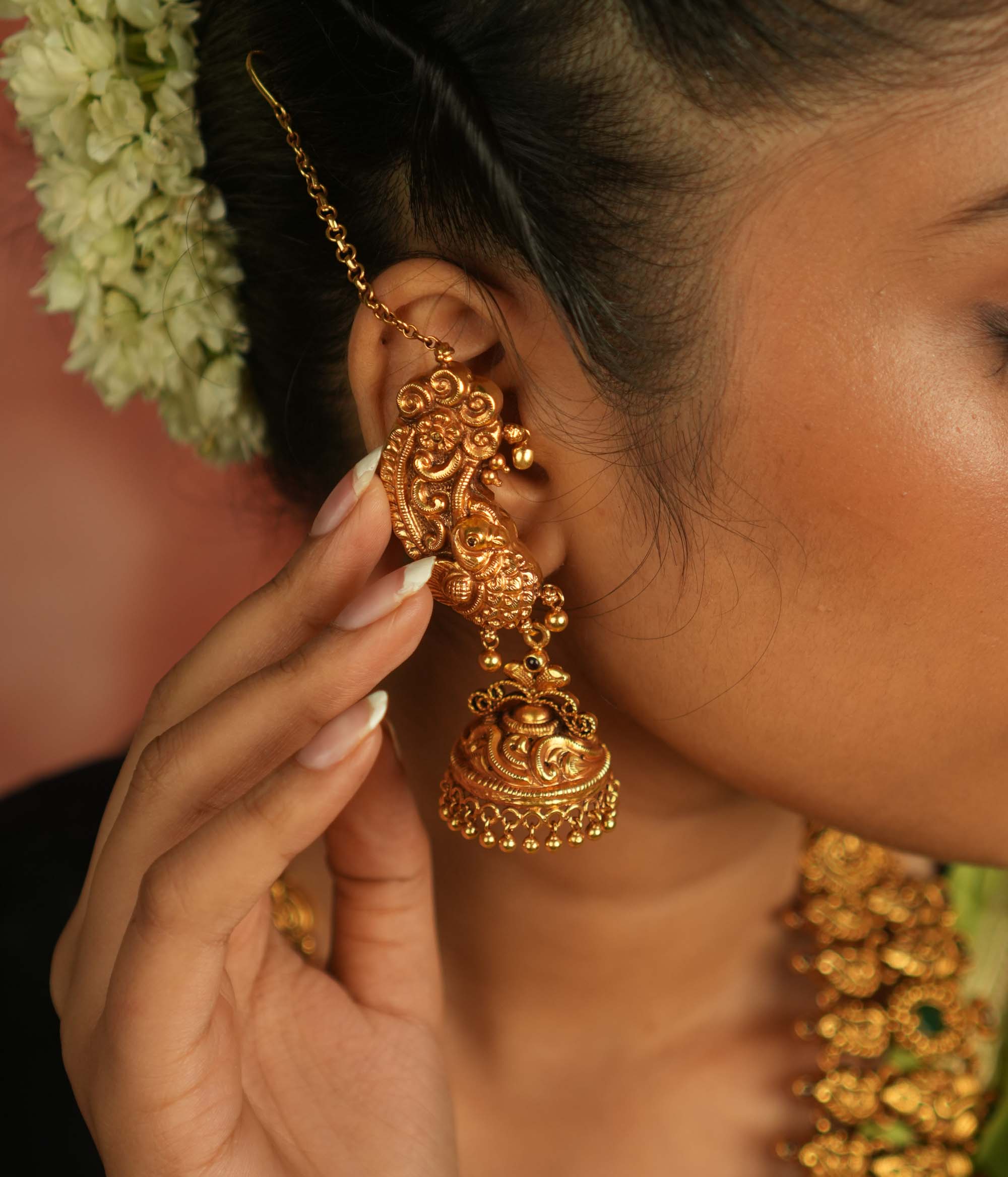 Regal peacock reborn! This gold-plated temple jhumka features intricate peacock motifs and a vibrant red spinel stone, with a delicate ear chain of tiny golden beads. A timeless blend of avian artistry and cultural opulence.