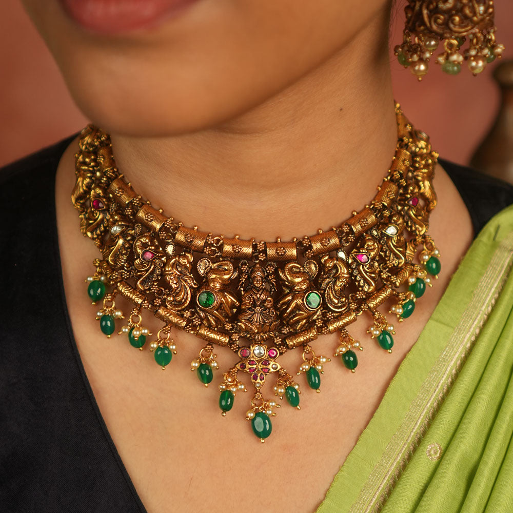 Temple whispers reborn! This antique gold necklace features Goddess Lakshmi flanked by intricate elephant and peacock motifs, adorned with vibrant red and green Kundan stones. Green beads and white pearl droplets add a celestial touch, celebrating mythical grandeur and cultural splendor.