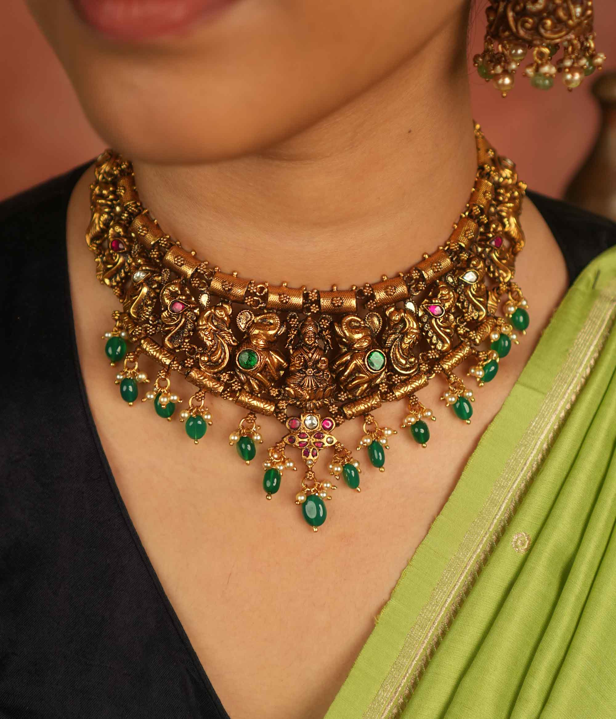 Temple whispers reborn! This antique gold necklace features Goddess Lakshmi flanked by intricate elephant and peacock motifs, adorned with vibrant red and green Kundan stones. Green beads and white pearl droplets add a celestial touch, celebrating mythical grandeur and cultural splendor.