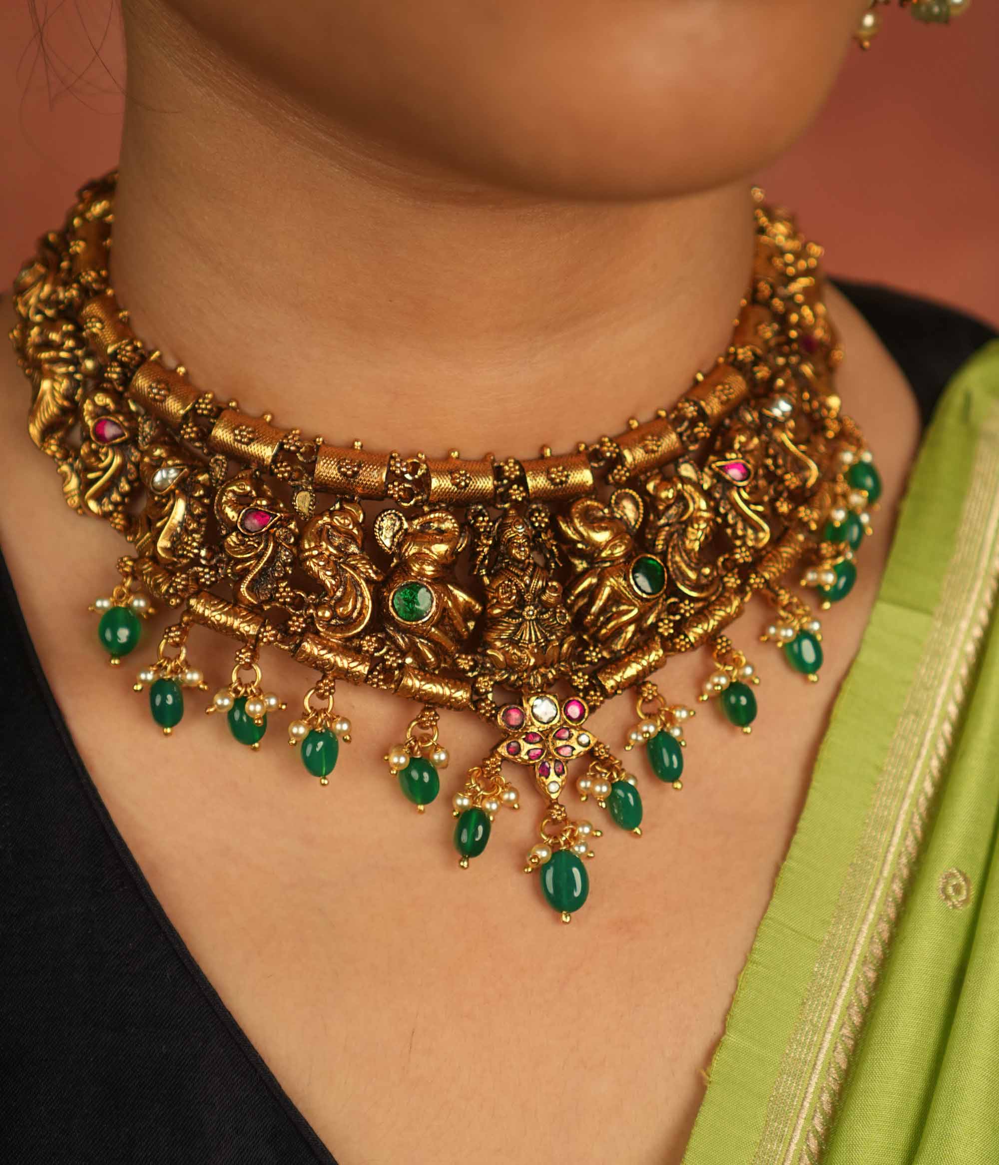 Temple whispers reborn! This antique gold necklace features Goddess Lakshmi flanked by intricate elephant and peacock motifs, adorned with vibrant red and green Kundan stones. Green beads and white pearl droplets add a celestial touch, celebrating mythical grandeur and cultural splendor.