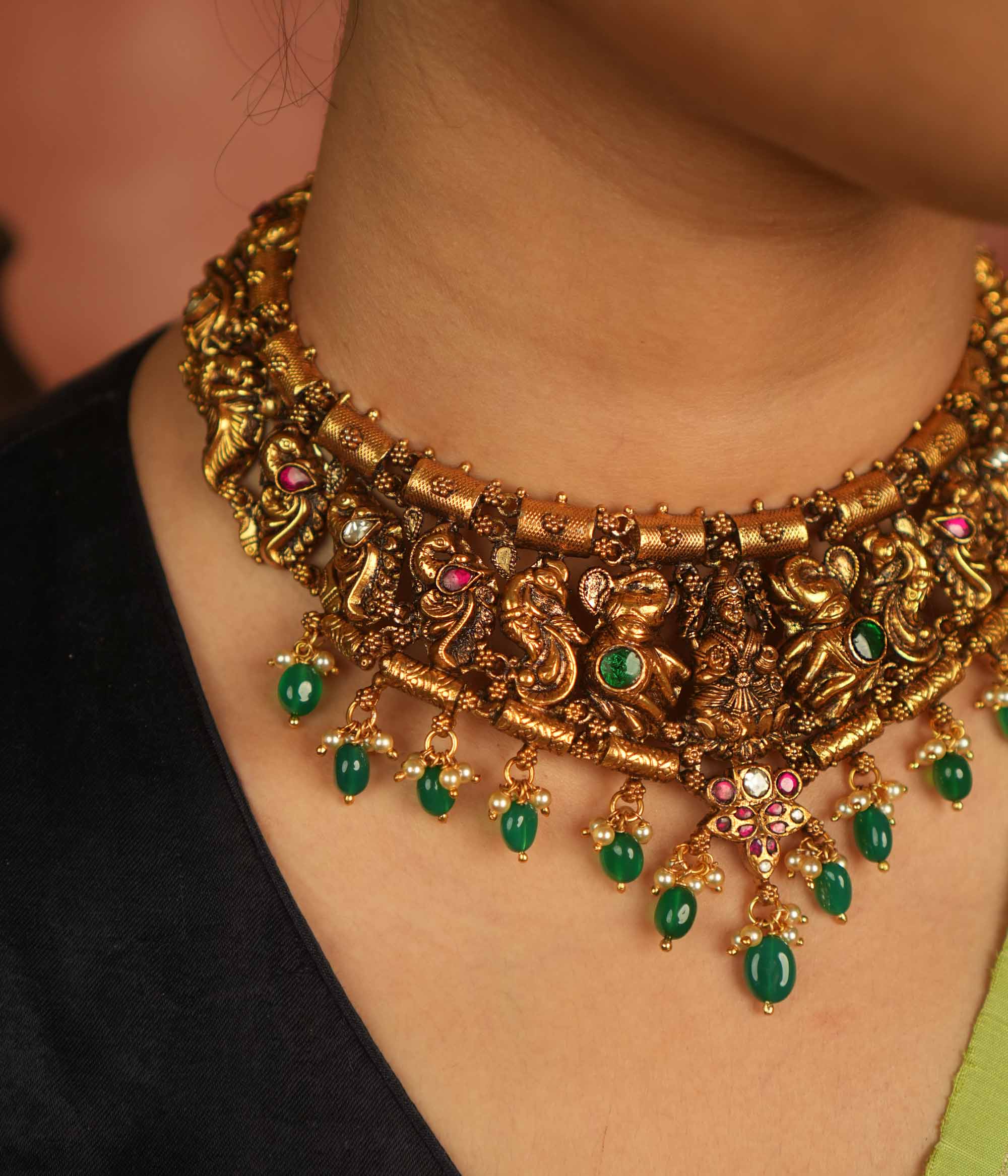 Temple whispers reborn! This antique gold necklace features Goddess Lakshmi flanked by intricate elephant and peacock motifs, adorned with vibrant red and green Kundan stones. Green beads and white pearl droplets add a celestial touch, celebrating mythical grandeur and cultural splendor.