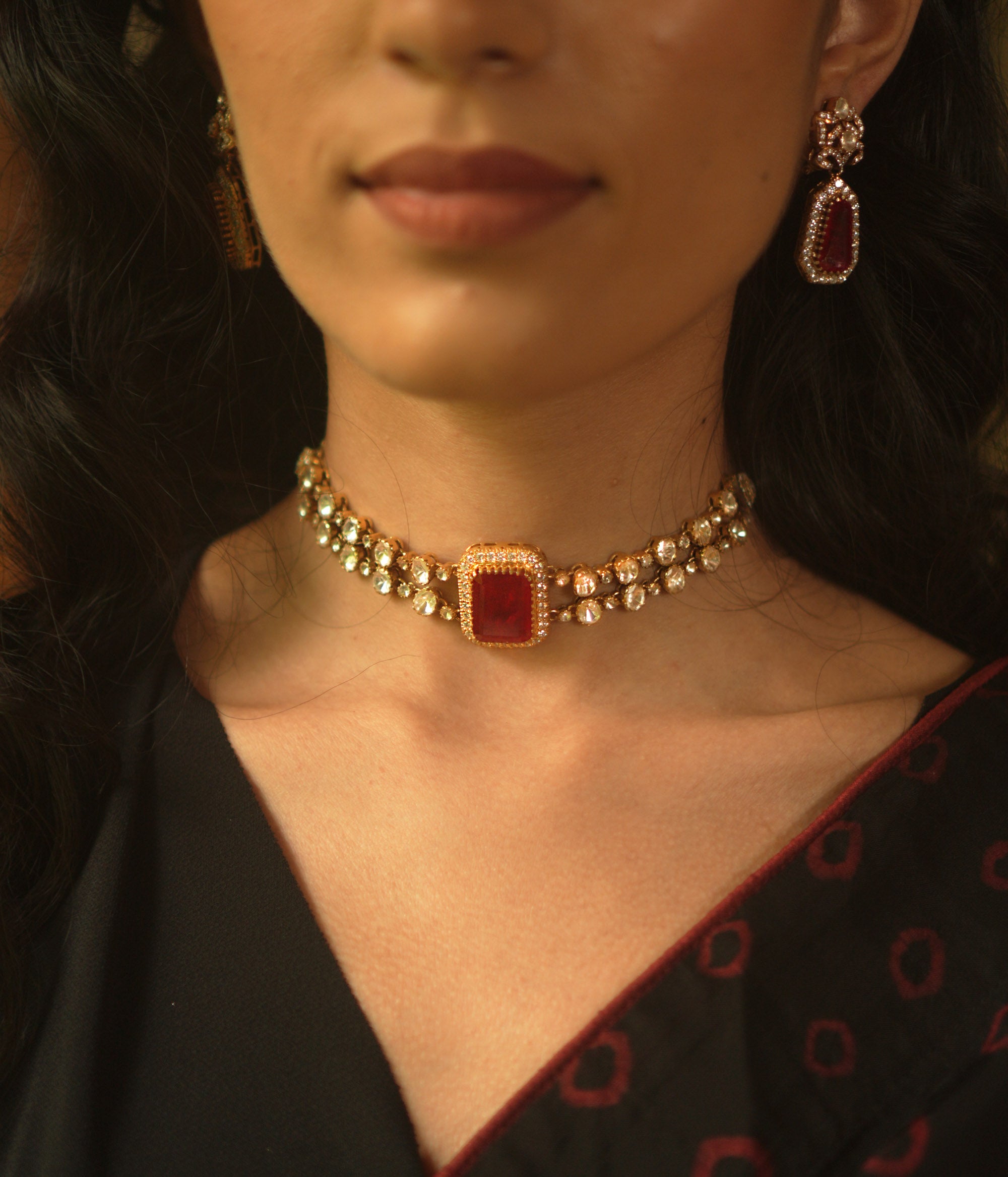 A gold choker with a central rectangular crimson stone, flanked by two rows of small clear stones, worn on a person's neck.