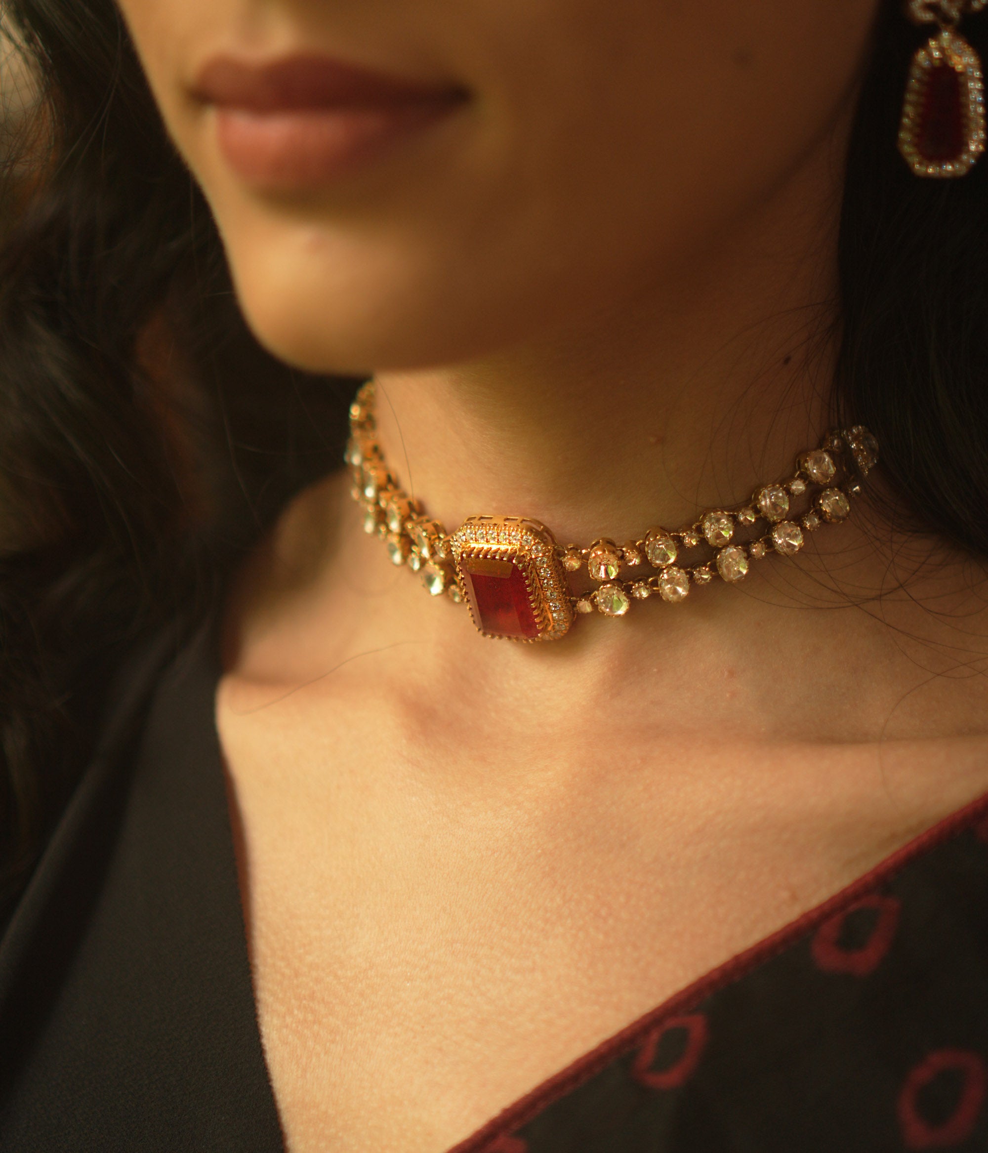 A gold choker with a central rectangular crimson stone, flanked by two rows of small clear stones, worn on a person's neck.