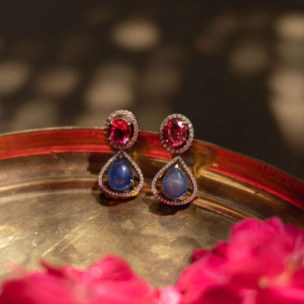 Close-up of Victorian-style teardrop earrings. Featuring pink and blue gemstones with diamond halos, resting on a textured gold surface with floral accents.