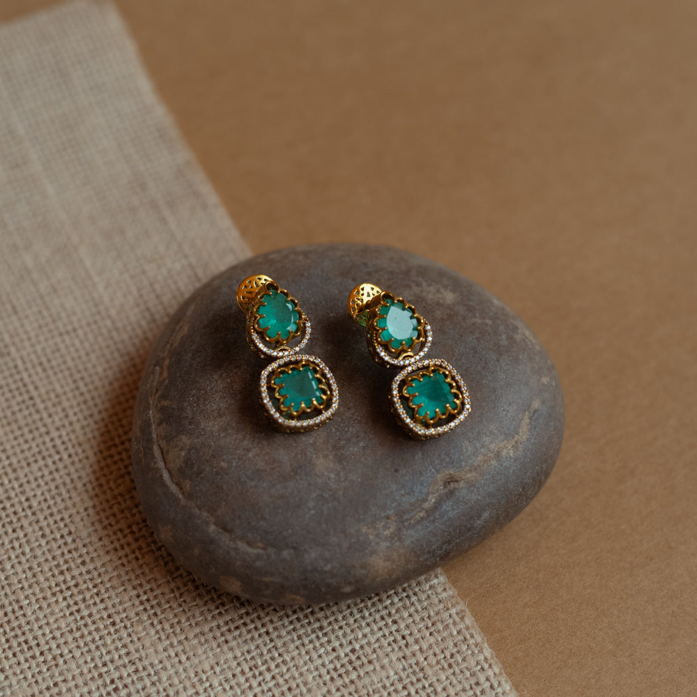 Gold drop earrings with green gemstones surrounded by CZ stones, worn by a woman.