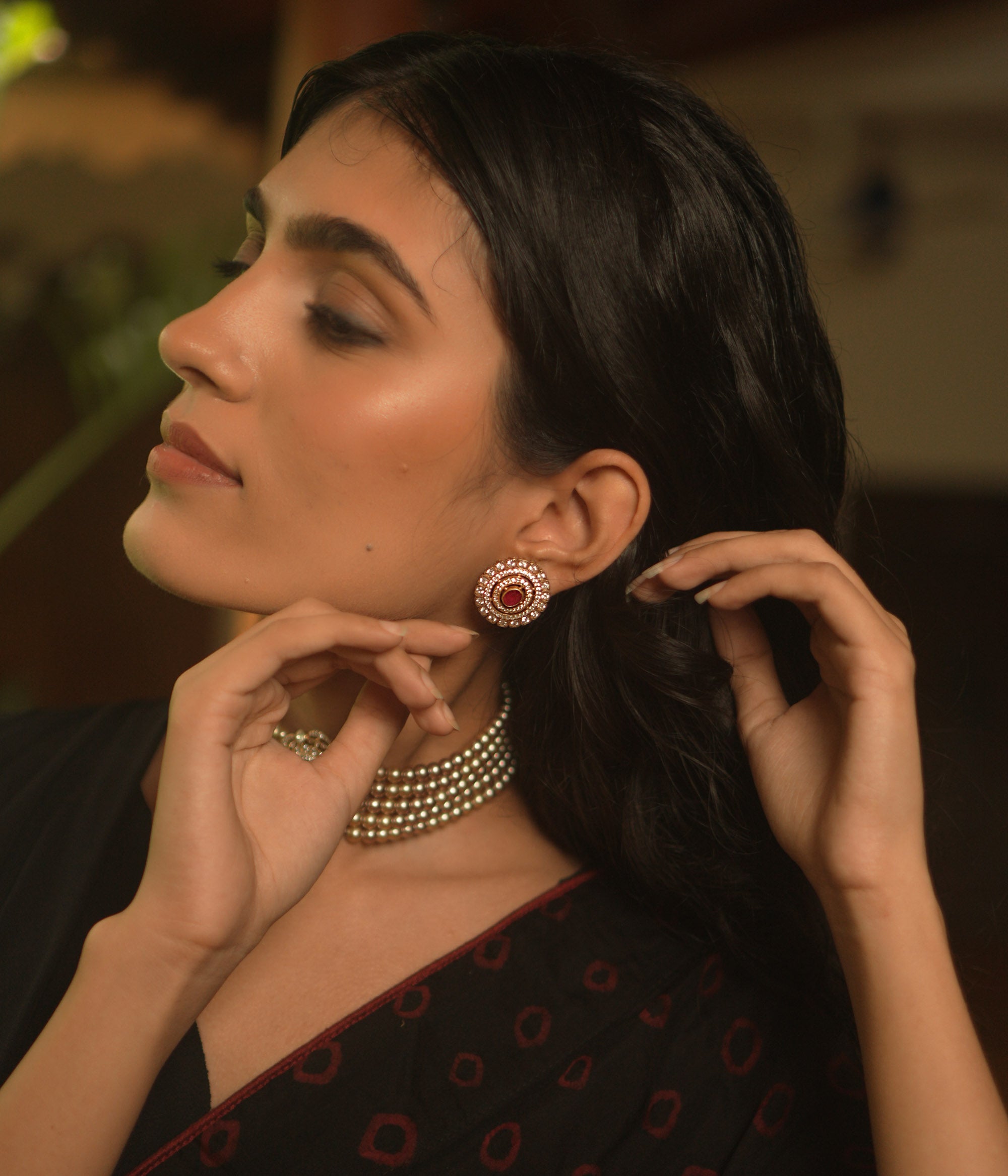 A pair of round stud earrings on a woman's ear, featuring a central red stone surrounded by concentric circles of white stones.