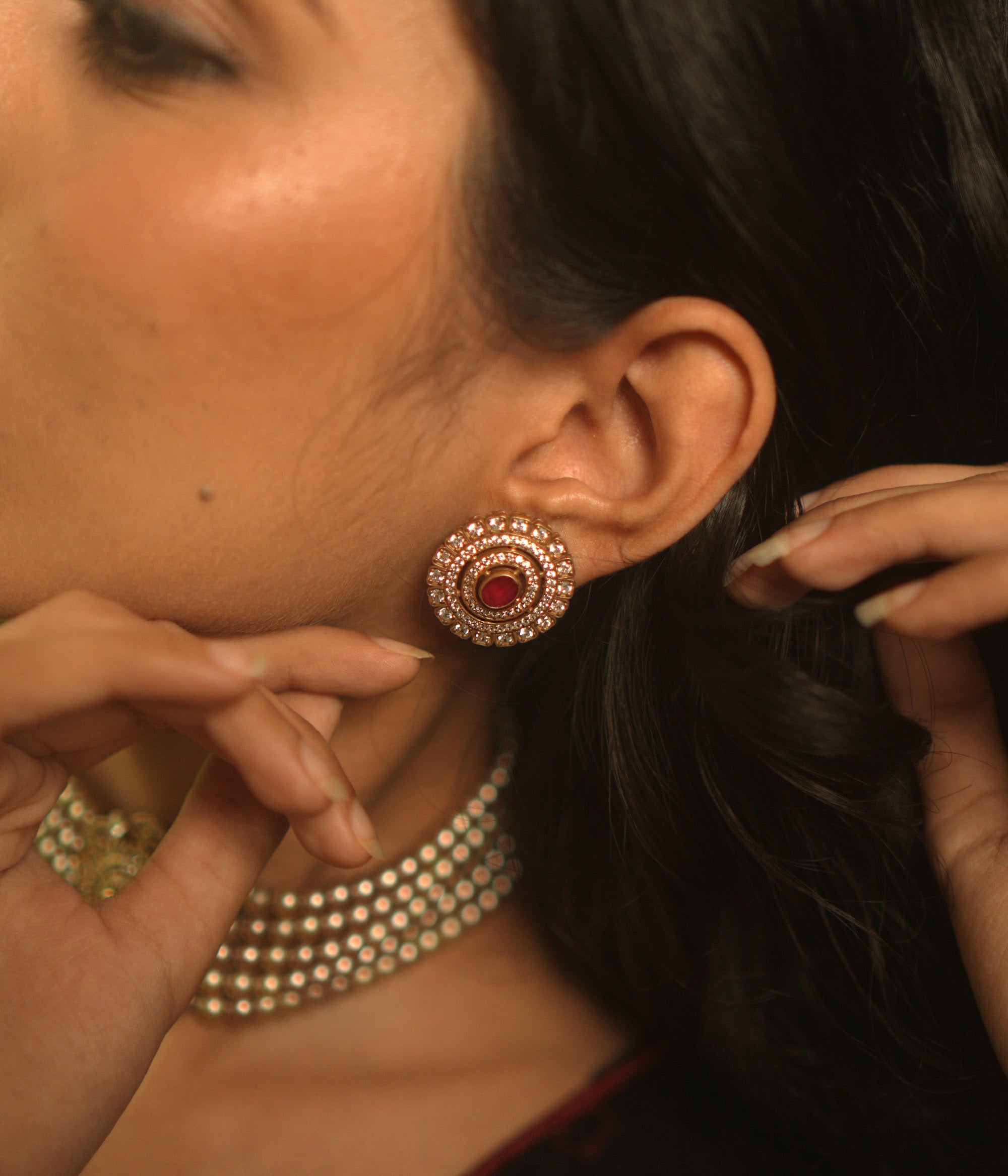 A pair of round stud earrings on a woman's ear, featuring a central red stone surrounded by concentric circles of white stones.