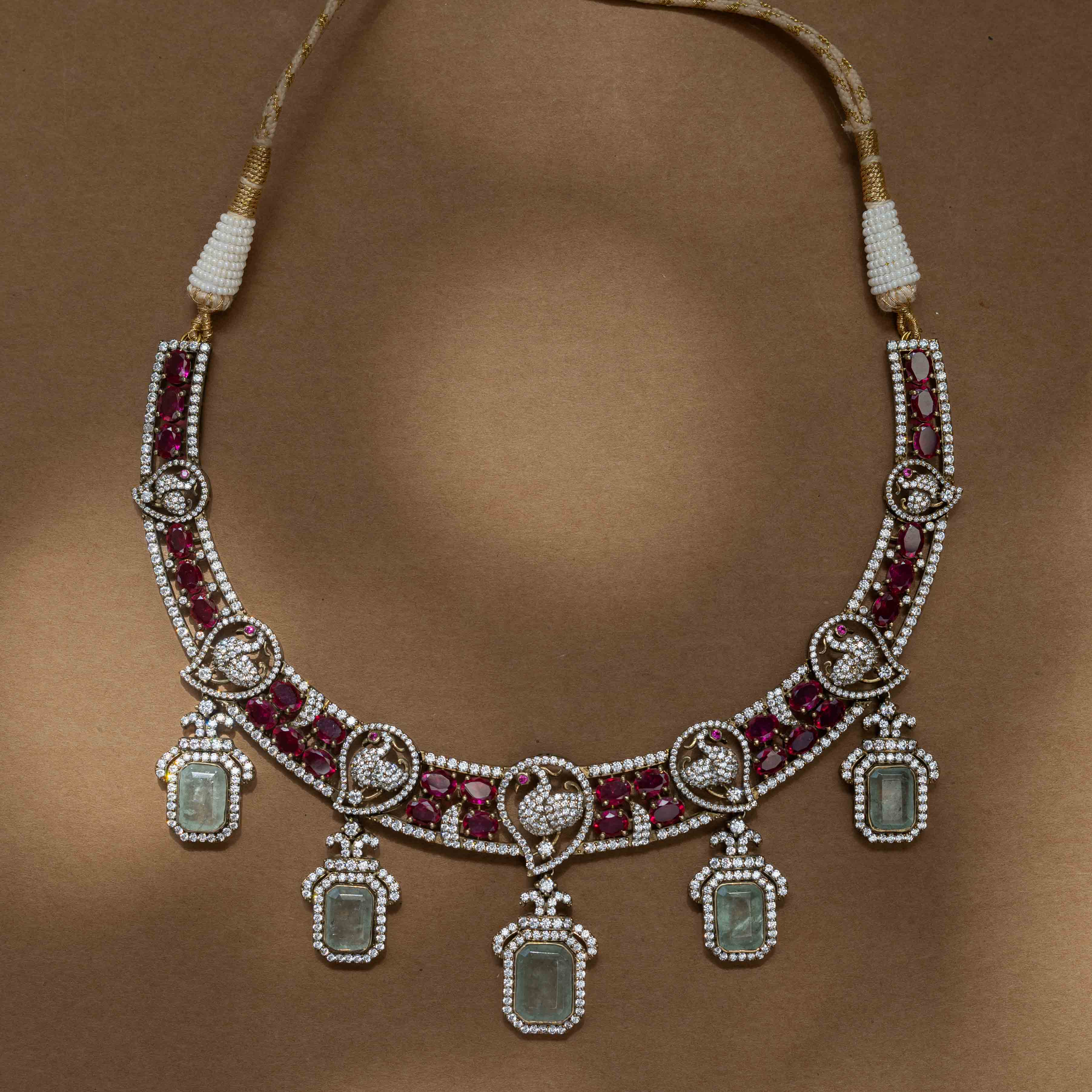 A two-tiered necklace with a large pendant featuring a green gem, clear and red stones, and pearl drops.