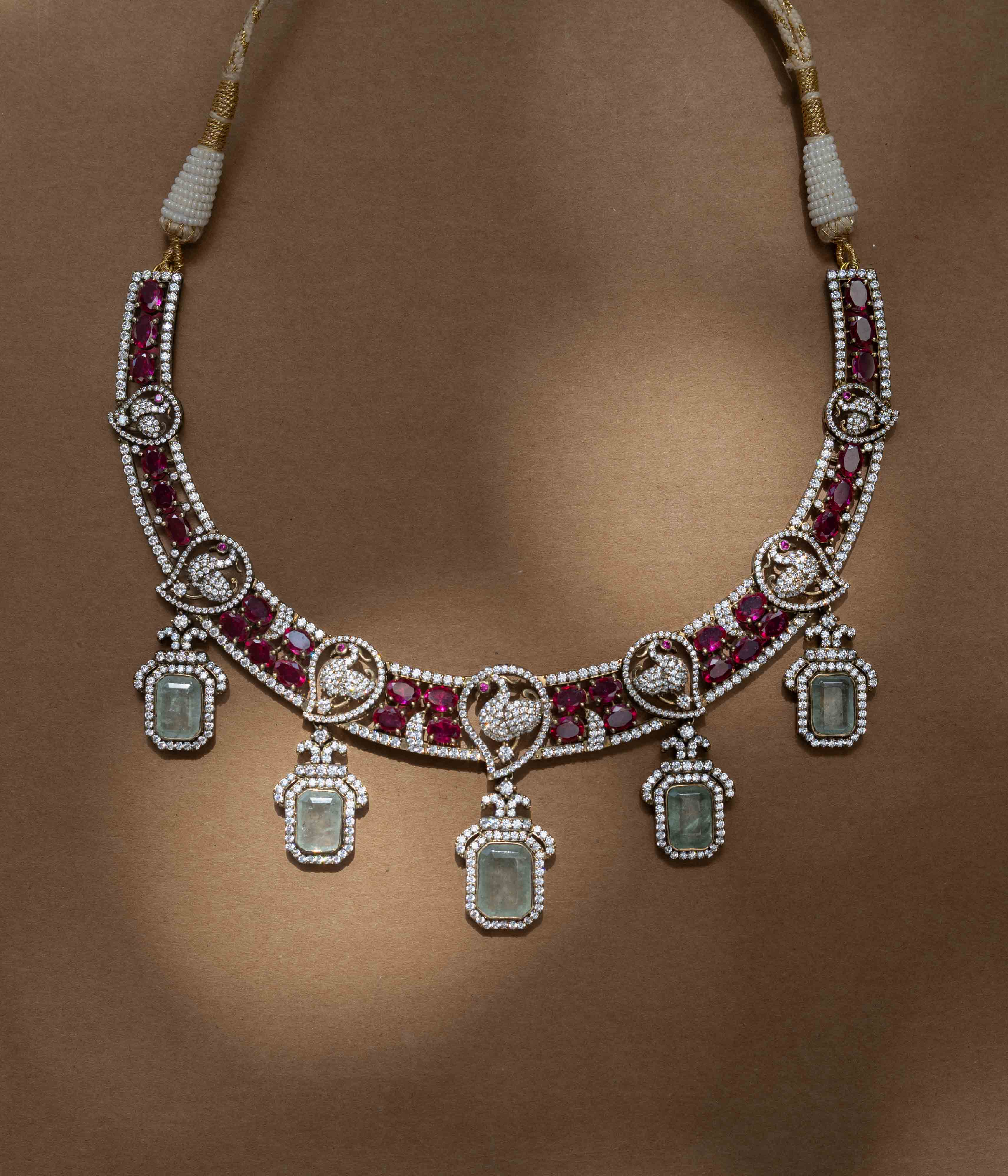 A two-tiered necklace with a large pendant featuring a green gem, clear and red stones, and pearl drops.