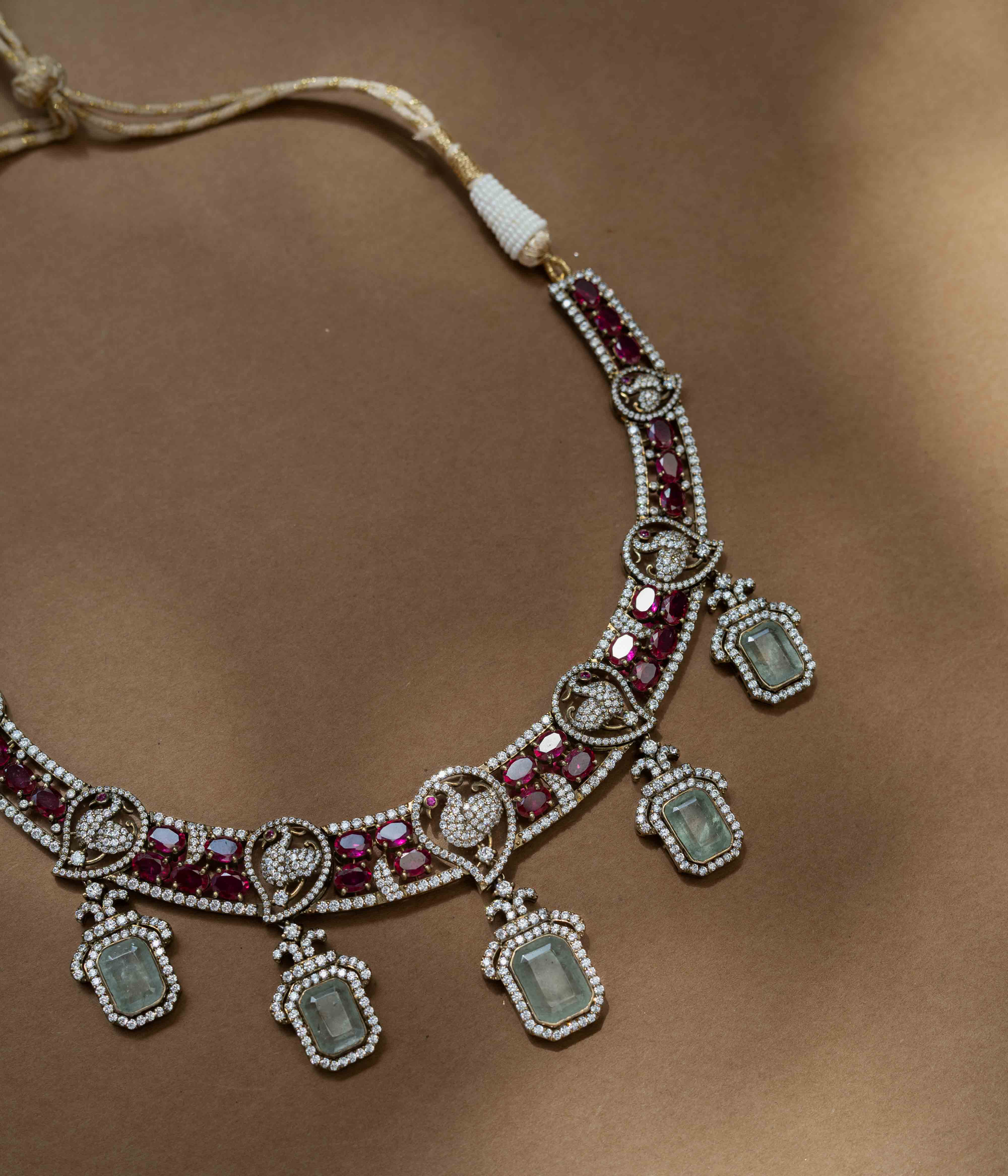 A two-tiered necklace with a large pendant featuring a green gem, clear and red stones, and pearl drops.