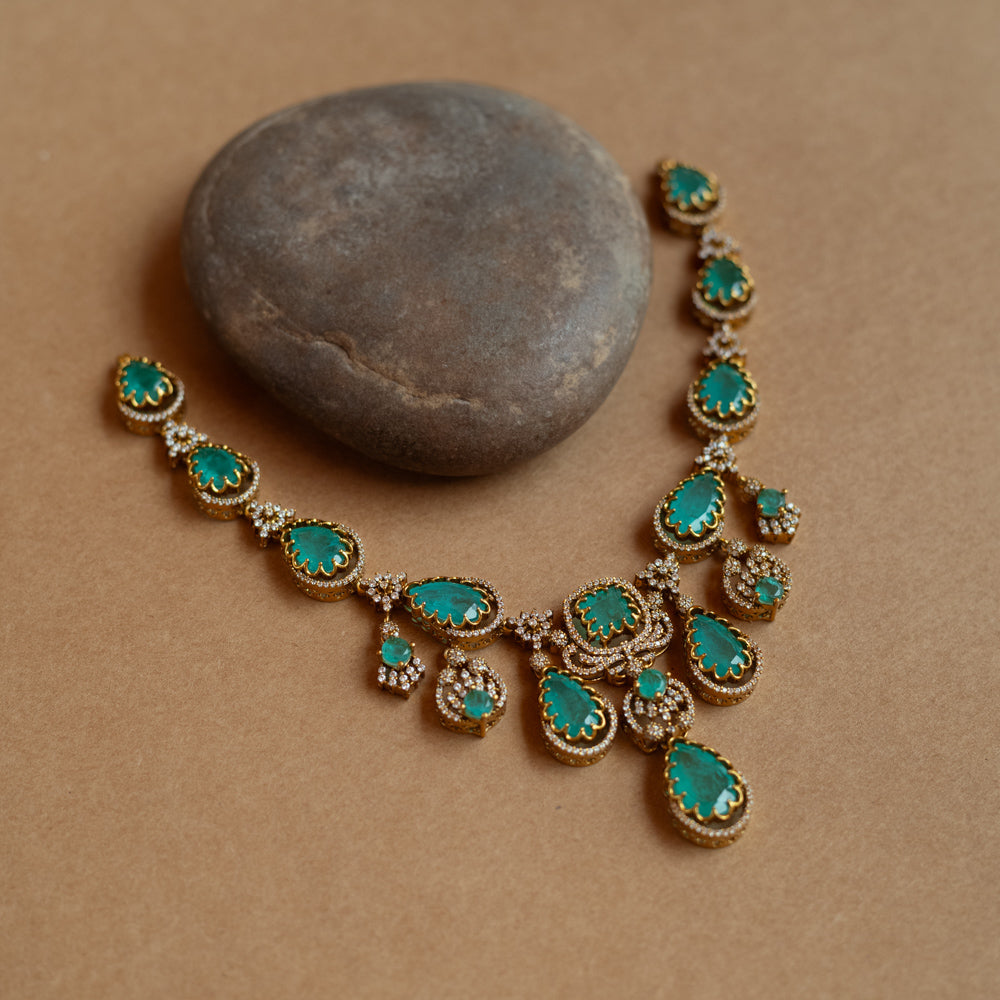 Gold necklace with large teardrop green gemstones surrounded by CZ stones, on a rock surface.