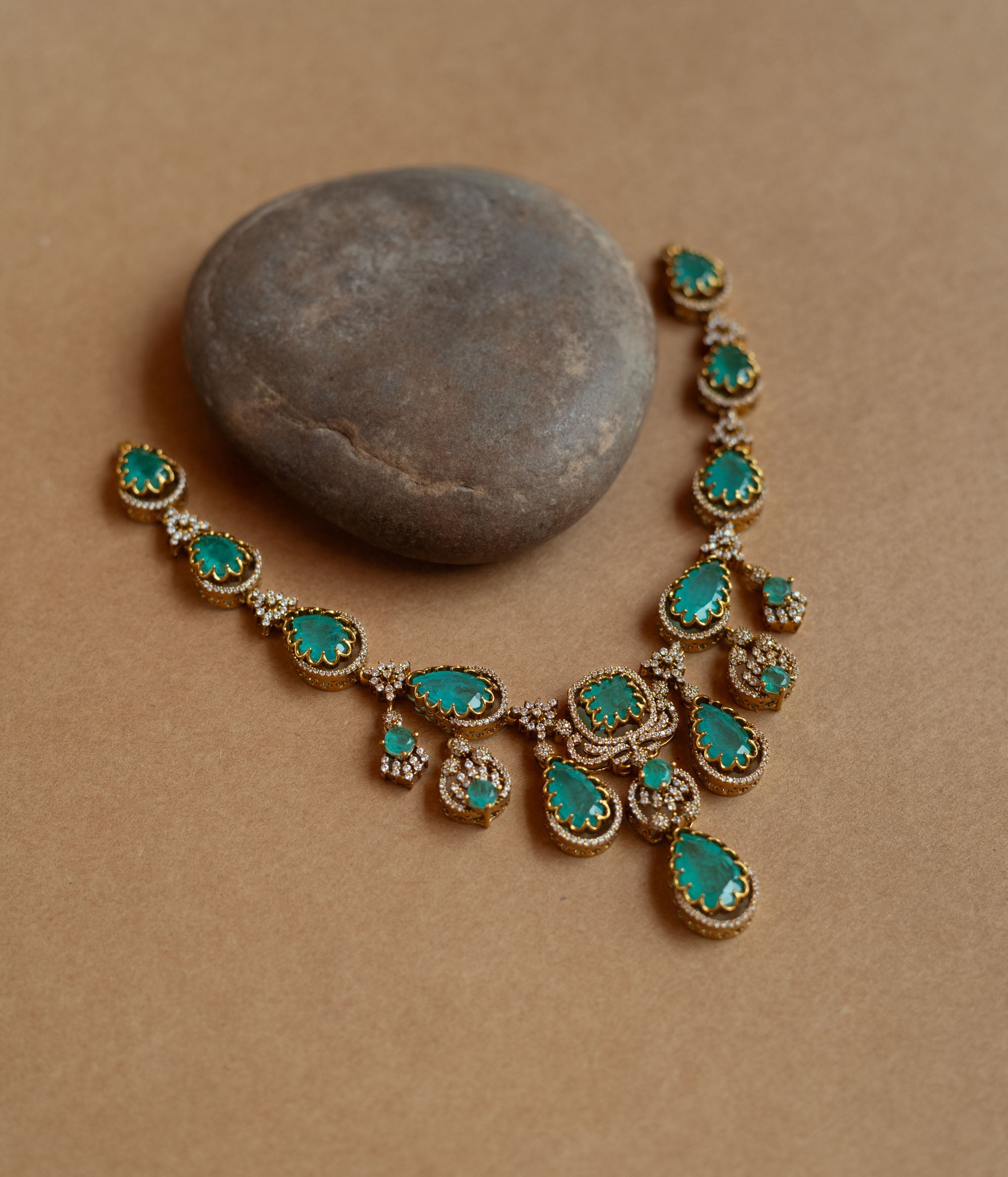 Gold necklace with large teardrop green gemstones surrounded by CZ stones, on a rock surface.