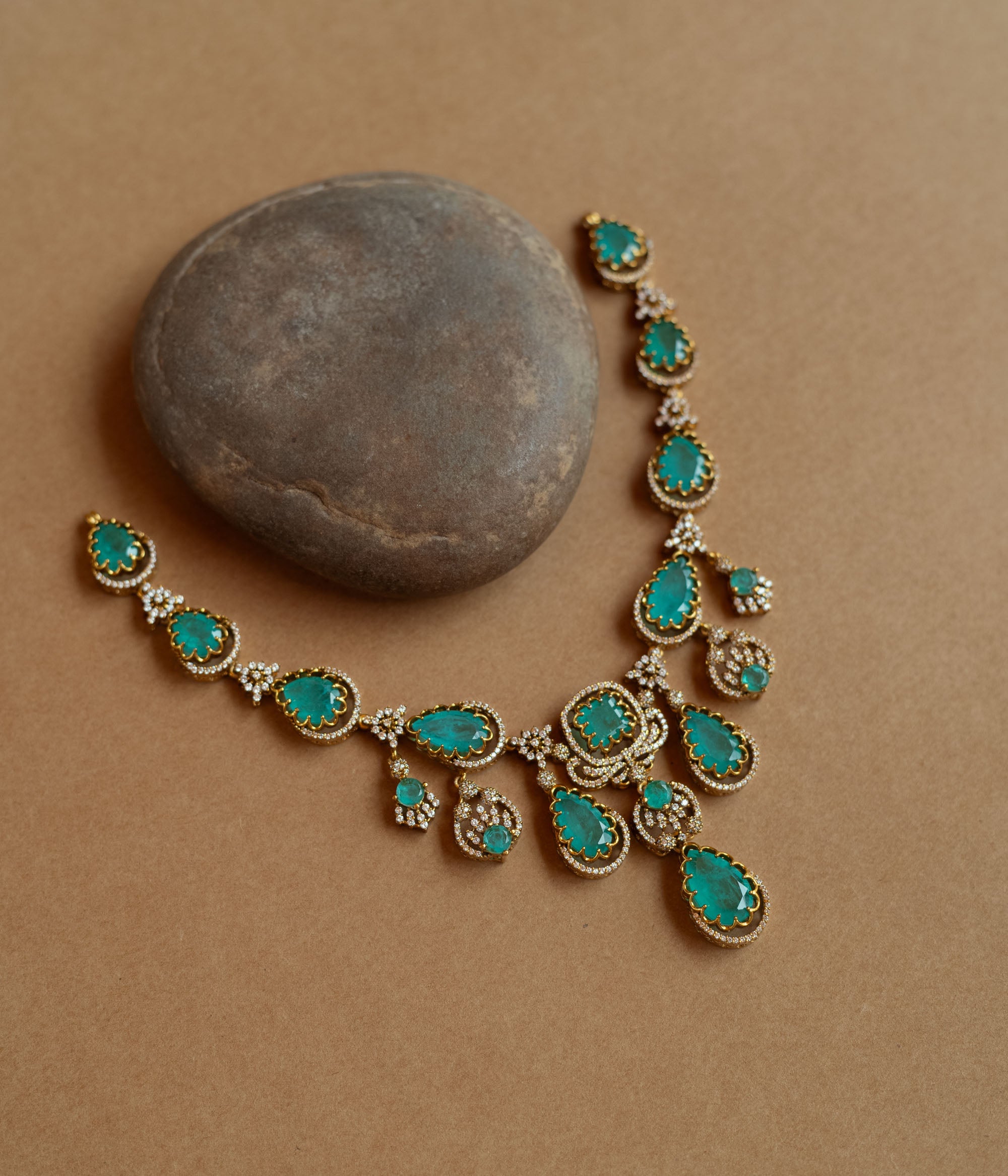 Gold necklace with large teardrop green gemstones surrounded by CZ stones, on a rock surface.