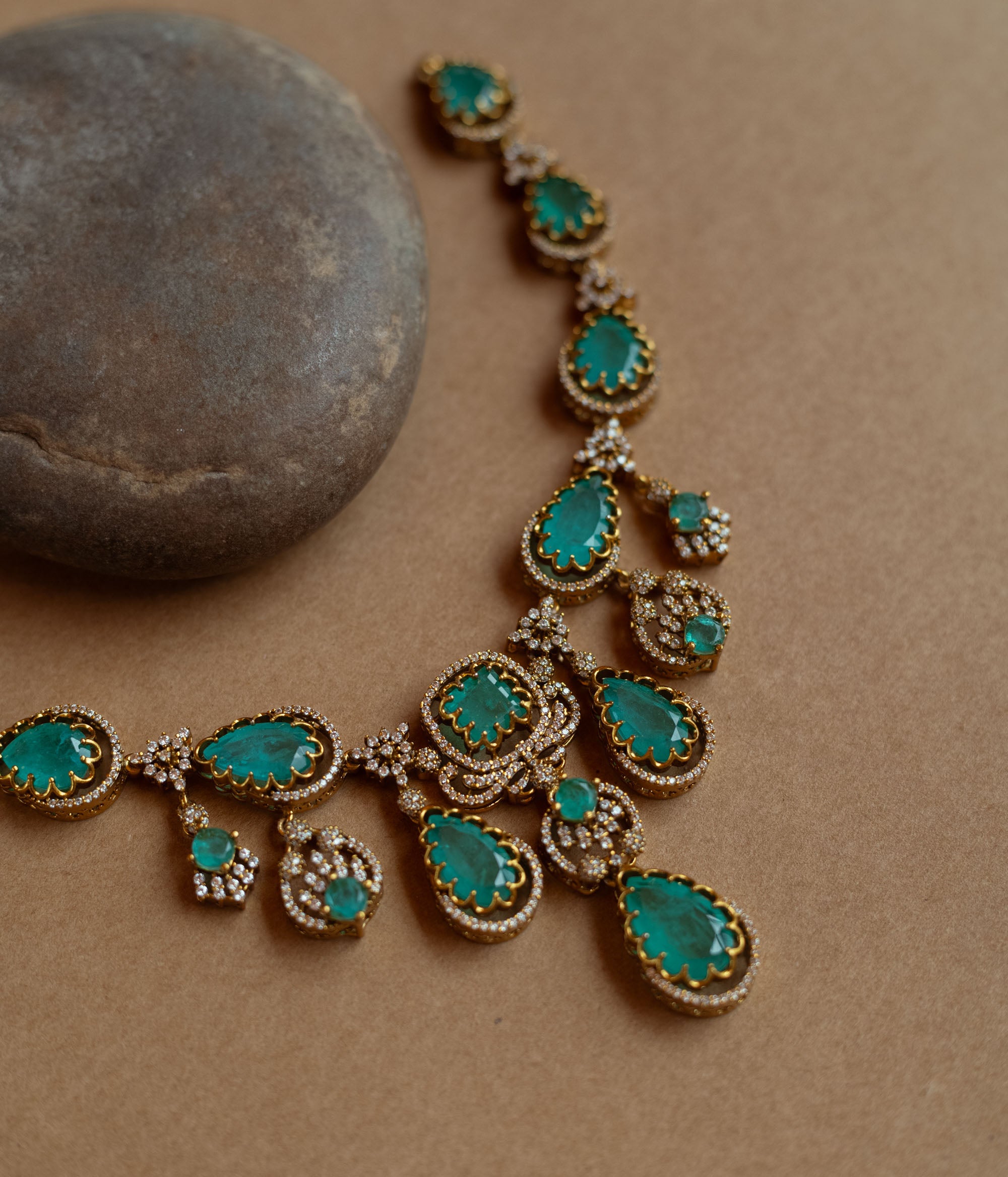 Gold necklace with large teardrop green gemstones surrounded by CZ stones, on a rock surface.