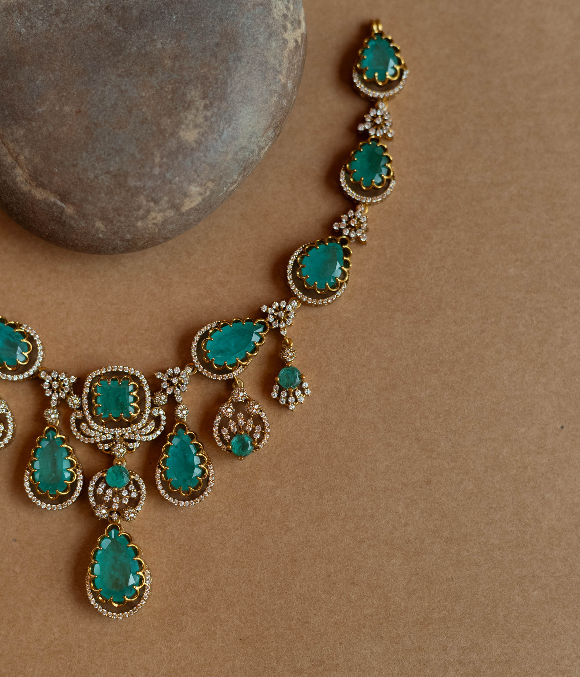 Gold necklace with large teardrop green gemstones surrounded by CZ stones, on a rock surface.
