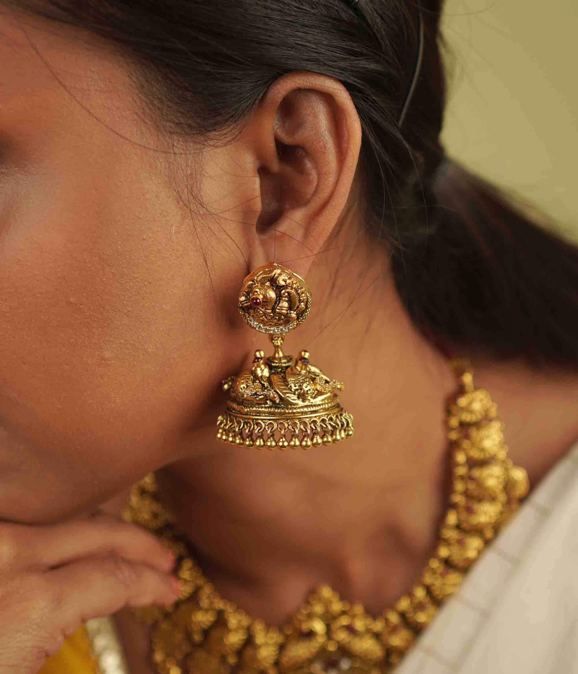 Regal grandeur meets timeless artistry! These gold-plated silver jhumkas, adorned with temple and peacock motifs, shimmer with red spinel and CZ stones, blending heritage craftsmanship with contemporary elegance.