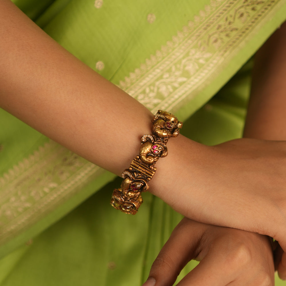 Embrace royal elegance with this stunning antique gold bangle, adorned with vibrant red, white, and green Kundan stones, featuring an intricate peacock motif. The regal peacock comes to life in this piece, while the pink Kundan hues add a touch of grandeur. A perfect fusion of heritage and artistry, this bangle exudes timeless splendor.