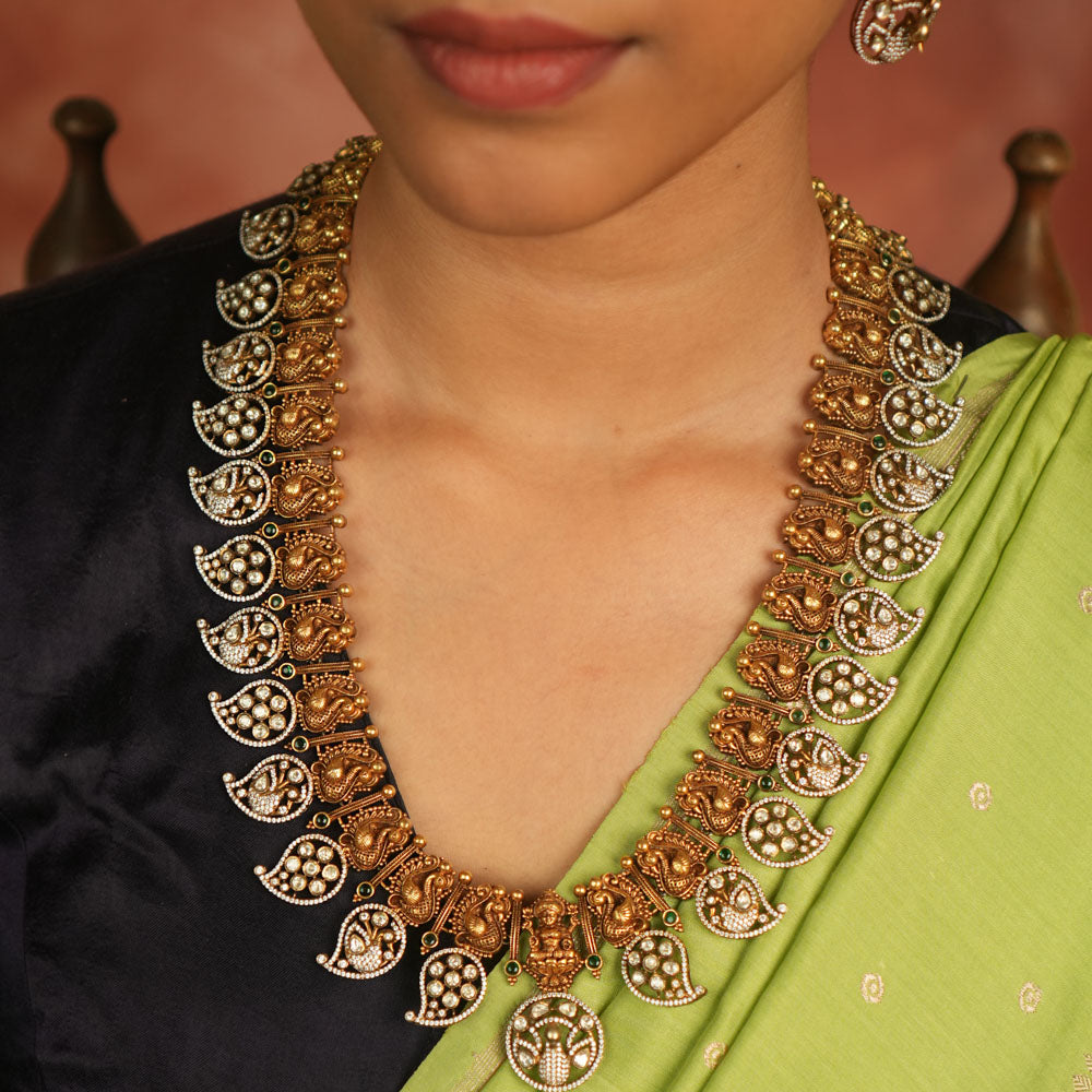 Temple whispers reborn! This gold-plated long necklace showcases regal peacock motifs and mango-shaped hangings adorned with polki, CZ, and green spinel stones. A timeless treasure that blends celestial grace with traditional charm.