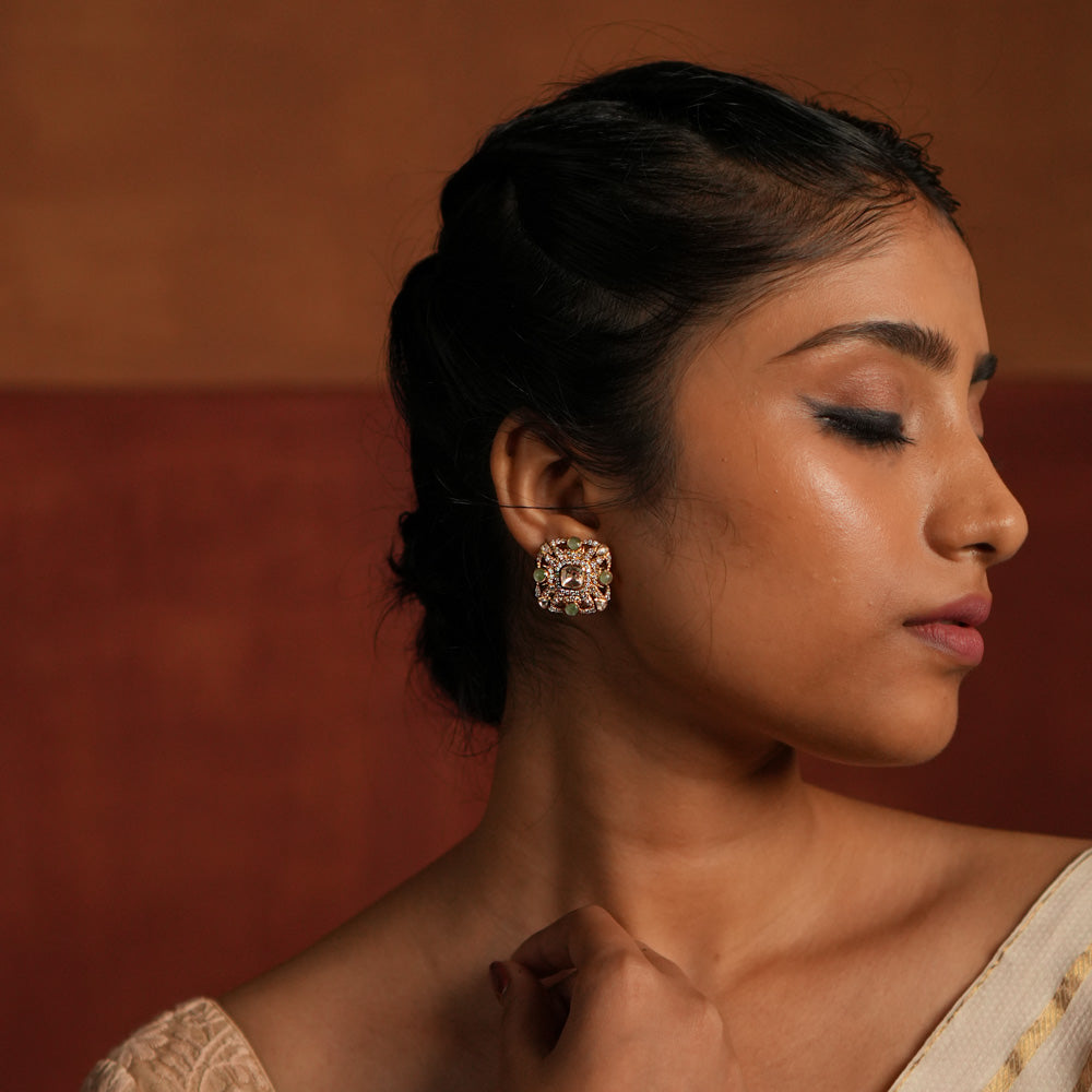 Elegant gold-plated silver stud earrings featuring a stunning square-shaped design. The center showcases a radiant Polki stone, beautifully encircled by layers of shimmering CZ stones. Adding to its charm, vibrant green gemstones are delicately placed within the CZ layers, enhancing the overall brilliance. A perfect blend of tradition and sophistication, ideal for any occasion.