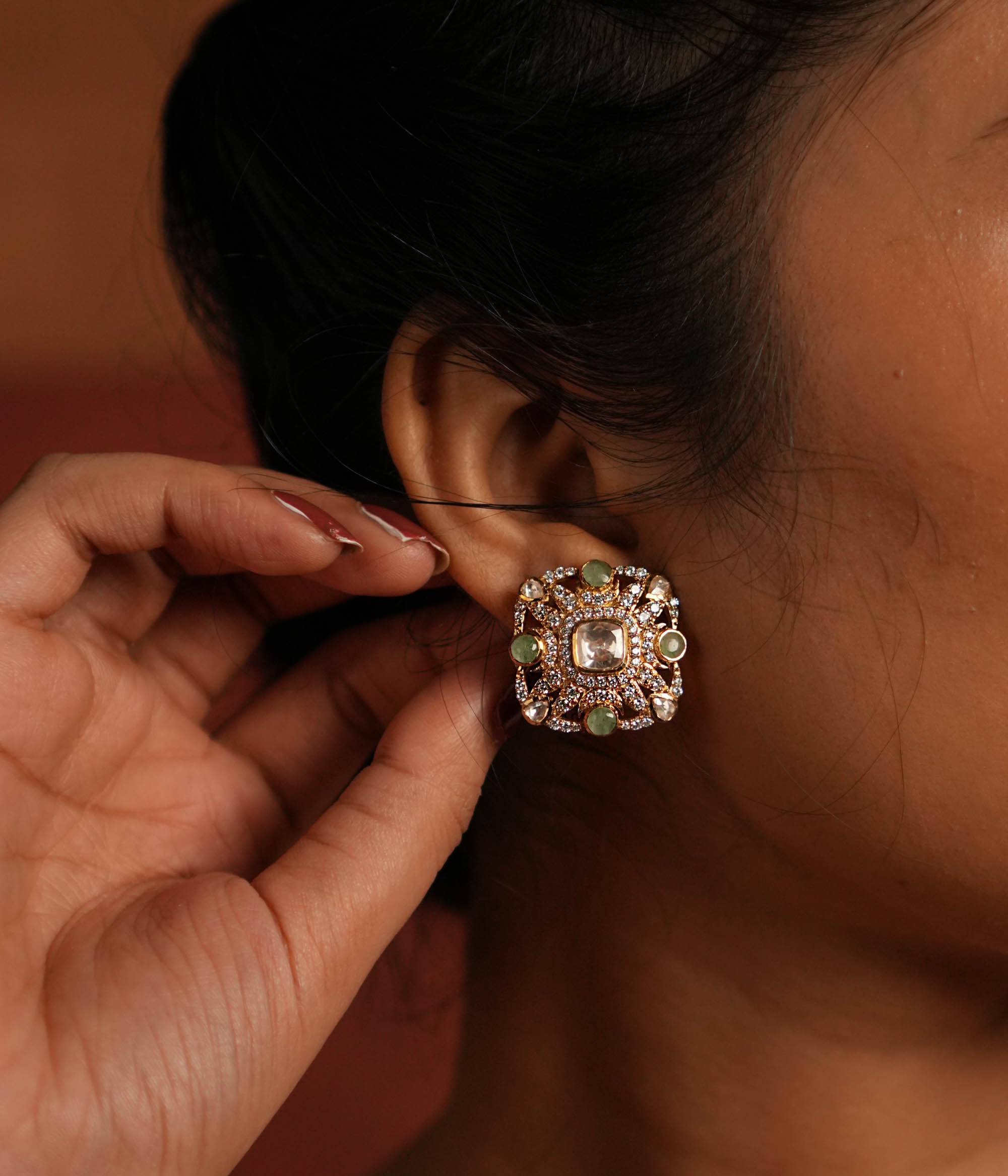 Elegant gold-plated silver stud earrings featuring a stunning square-shaped design. The center showcases a radiant Polki stone, beautifully encircled by layers of shimmering CZ stones. Adding to its charm, vibrant green gemstones are delicately placed within the CZ layers, enhancing the overall brilliance. A perfect blend of tradition and sophistication, ideal for any occasion.
