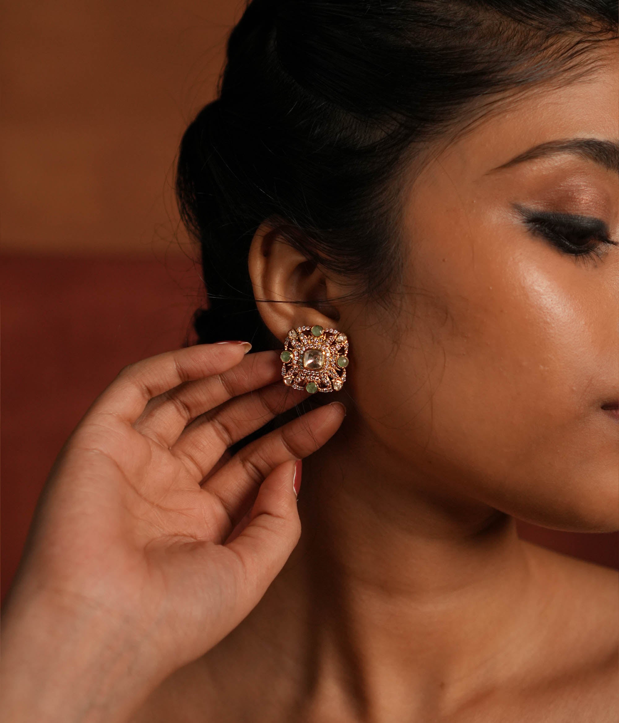 Elegant gold-plated silver stud earrings featuring a stunning square-shaped design. The center showcases a radiant Polki stone, beautifully encircled by layers of shimmering CZ stones. Adding to its charm, vibrant green gemstones are delicately placed within the CZ layers, enhancing the overall brilliance. A perfect blend of tradition and sophistication, ideal for any occasion.